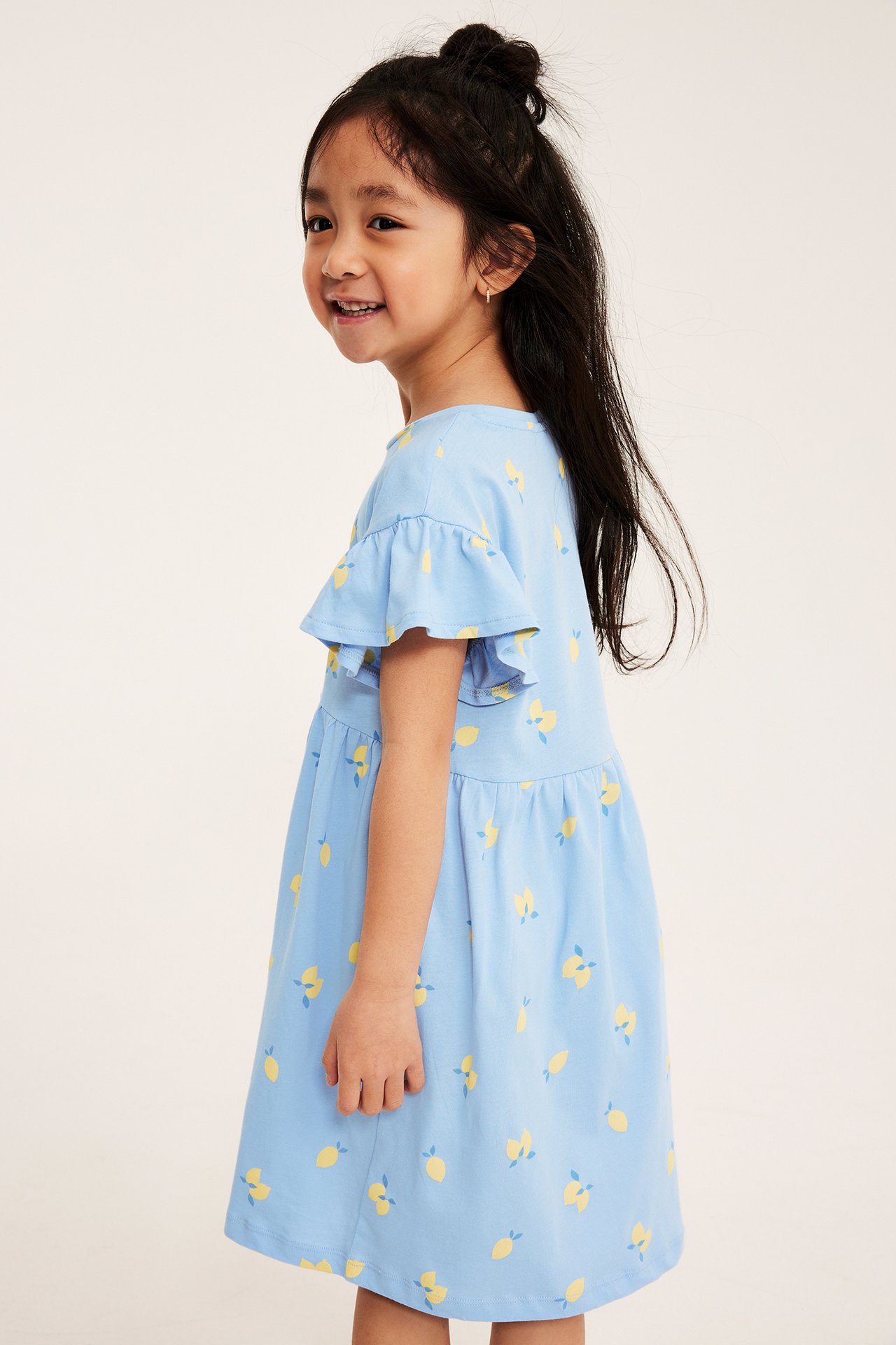 Widok z boku dziecka w jasnoniebieskiej bawełnianej sukience z falbanami i wzorem w cytryny.