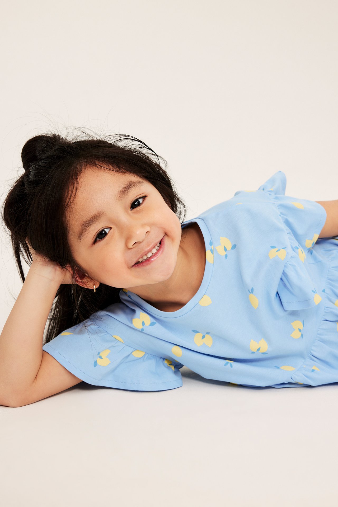 Widok z przodu uśmiechniętego dziecka w jasnoniebieskiej sukience bawełnianej w żółte cytryny, z falbankami.