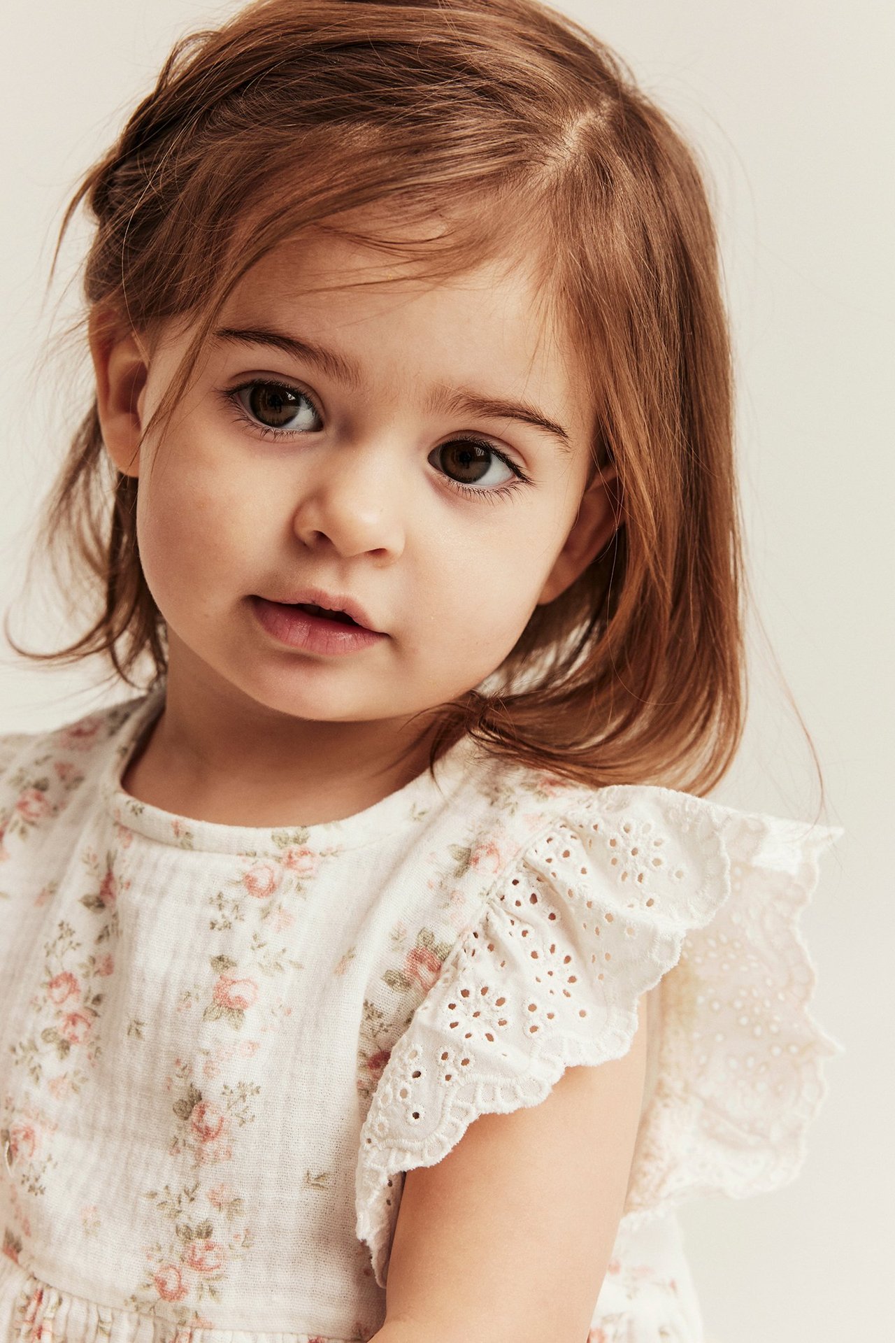 Nærbilde forfra av et barn i en naturhvit blomstret musselintopp med broderte rysjeermer.