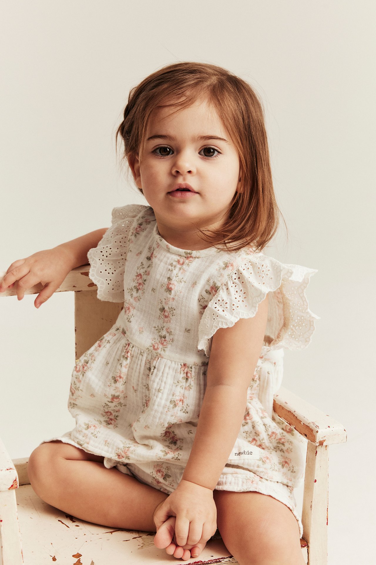 Framifrån, en baby sitter i ett offwhite blommigt muslin shorts-set med peplumtopp och broderade volangärmar.