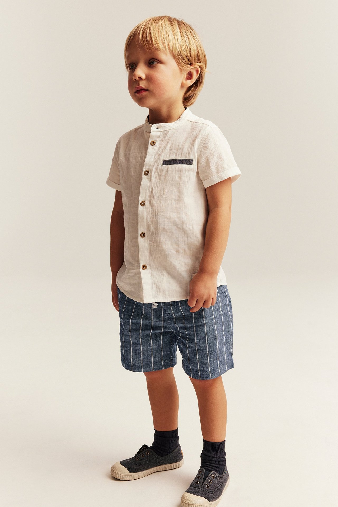 Forfra, et barn i blåstripete vevde shorts, en hvit linblandet skjorte, mørke sokker og grå seilersko.