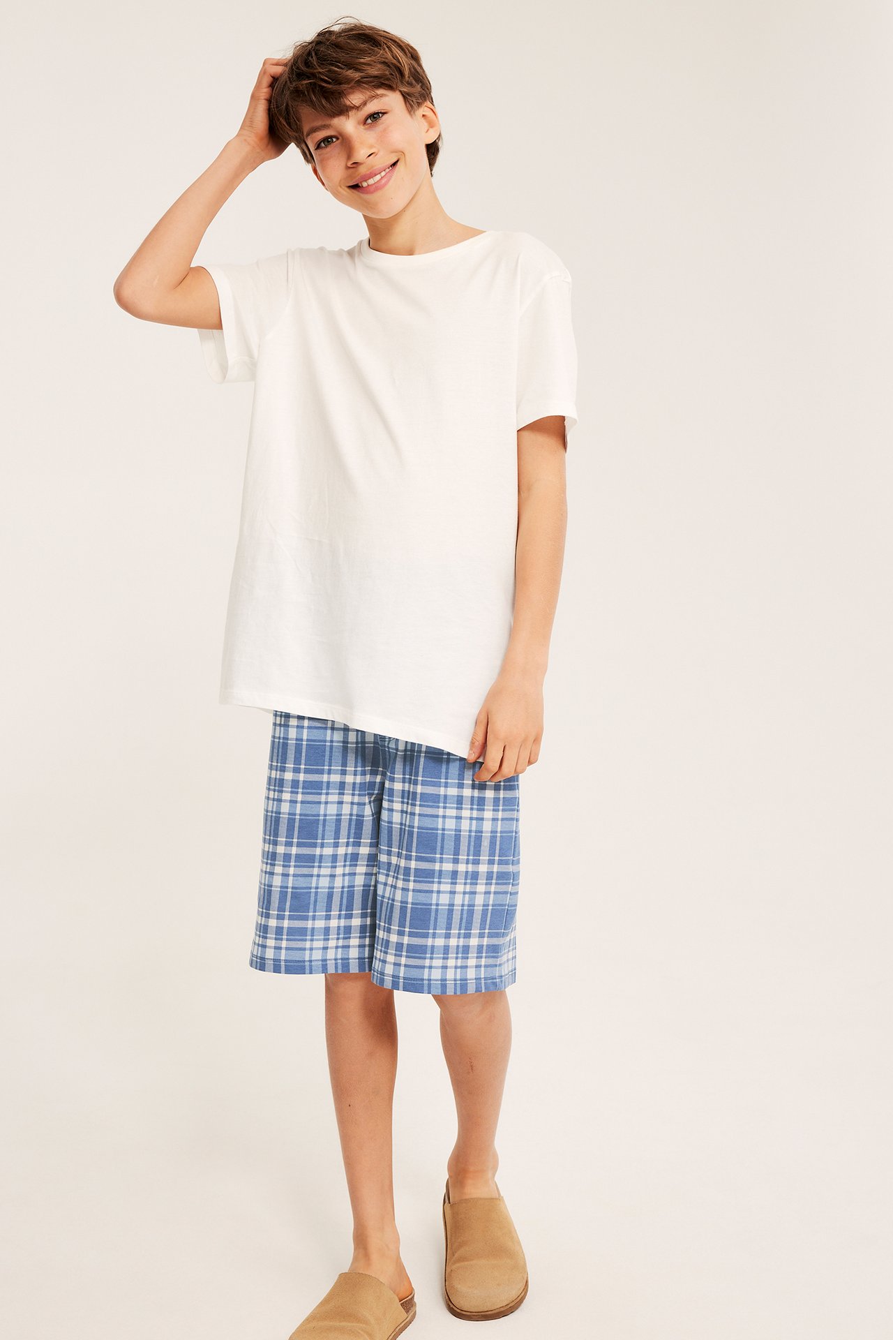 Framifrån, ett barn i blårutiga pyjamasshorts i bomull, en vit t-shirt och bruna tofflor.