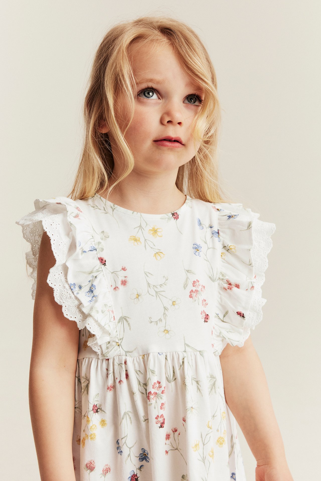 Nærbilde forfra av et barn i offwhite blomstret jerseykjole med rysjer og blondedetaljer.