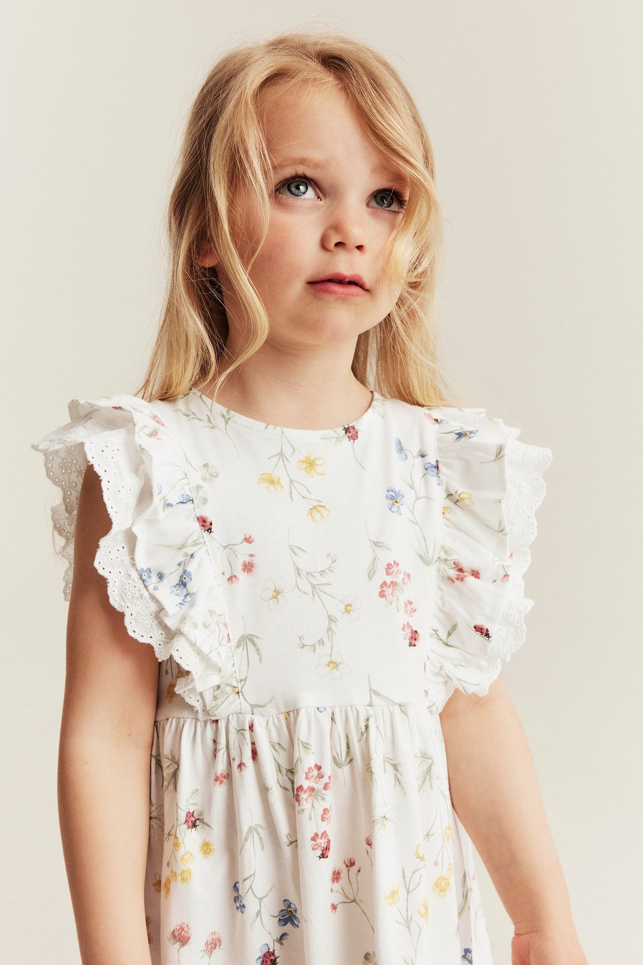 Lähikuva edestä lapsesta luonnonvalkoisessa kukkakuviollisessa trikoomekossa, jossa röyhelö- ja pitsihihat.
