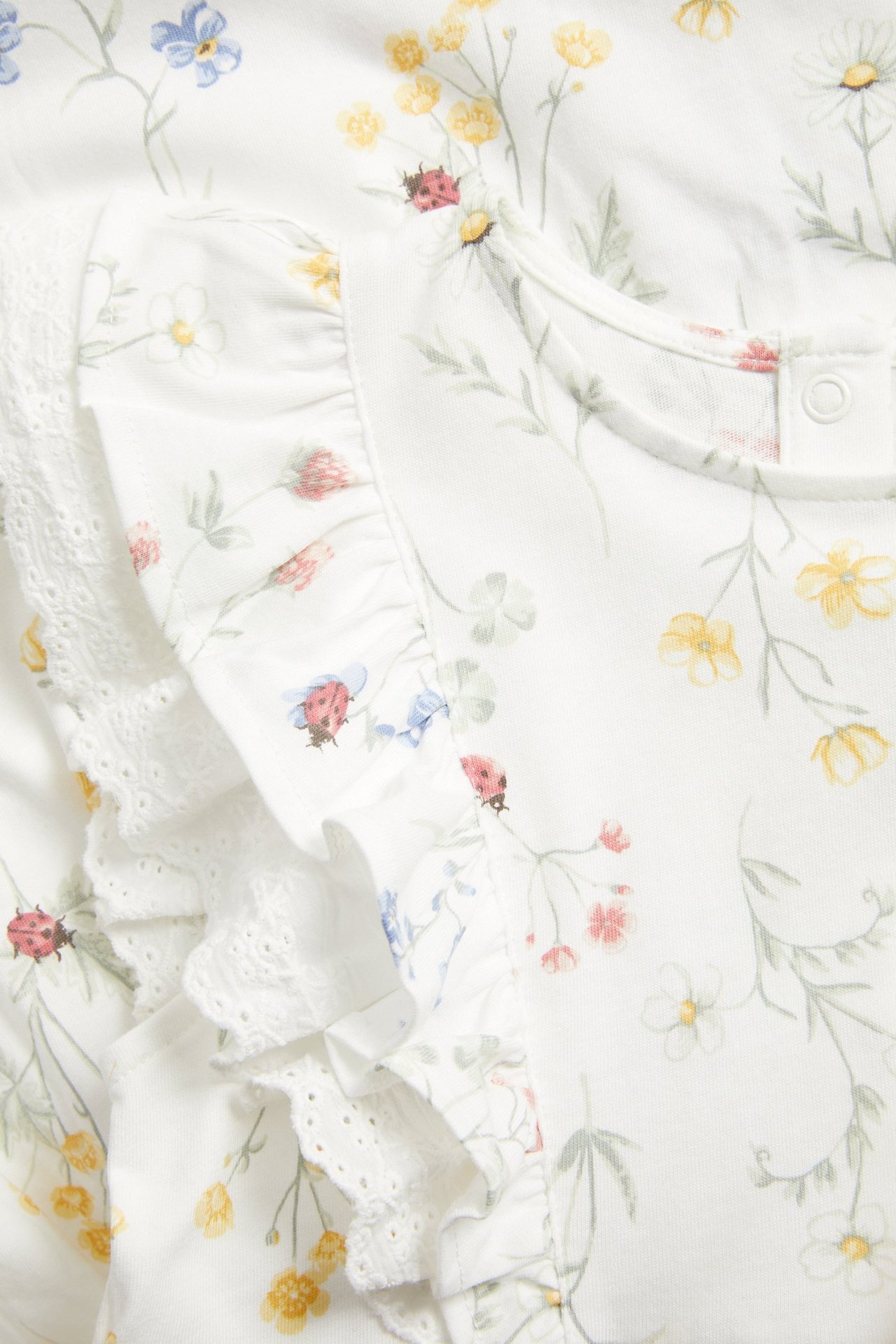 Nærbilde av en offwhite blomstret jerseykjole med volanger, blondedetaljer, trykknapper og marihønetrykk.