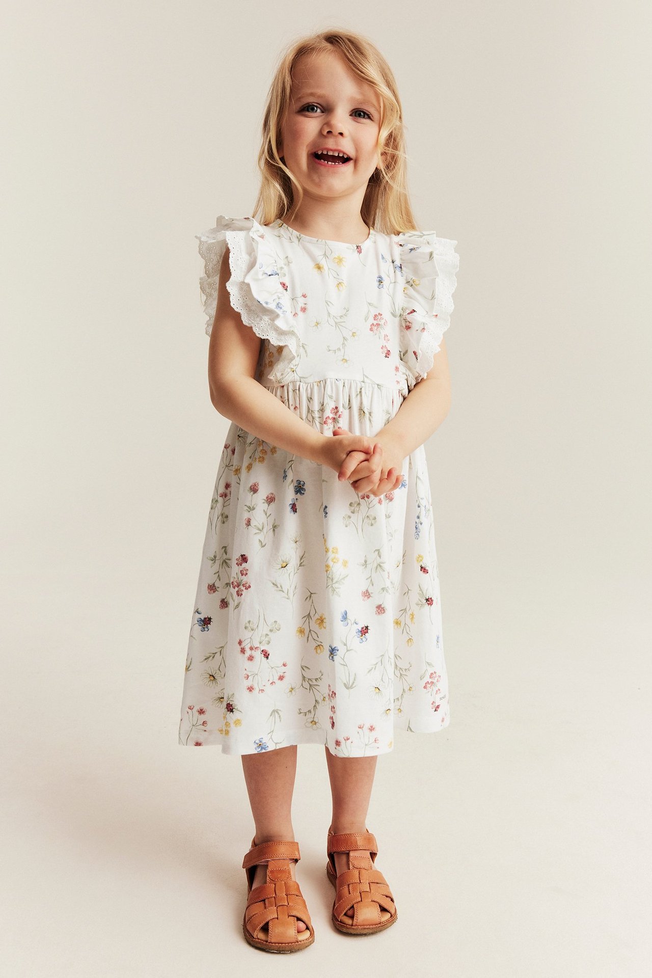 Widok z przodu: dziewczynka w ecru kwiecistej sukience z dżerseju z falbankami i jasnobrązowymi sandałami.