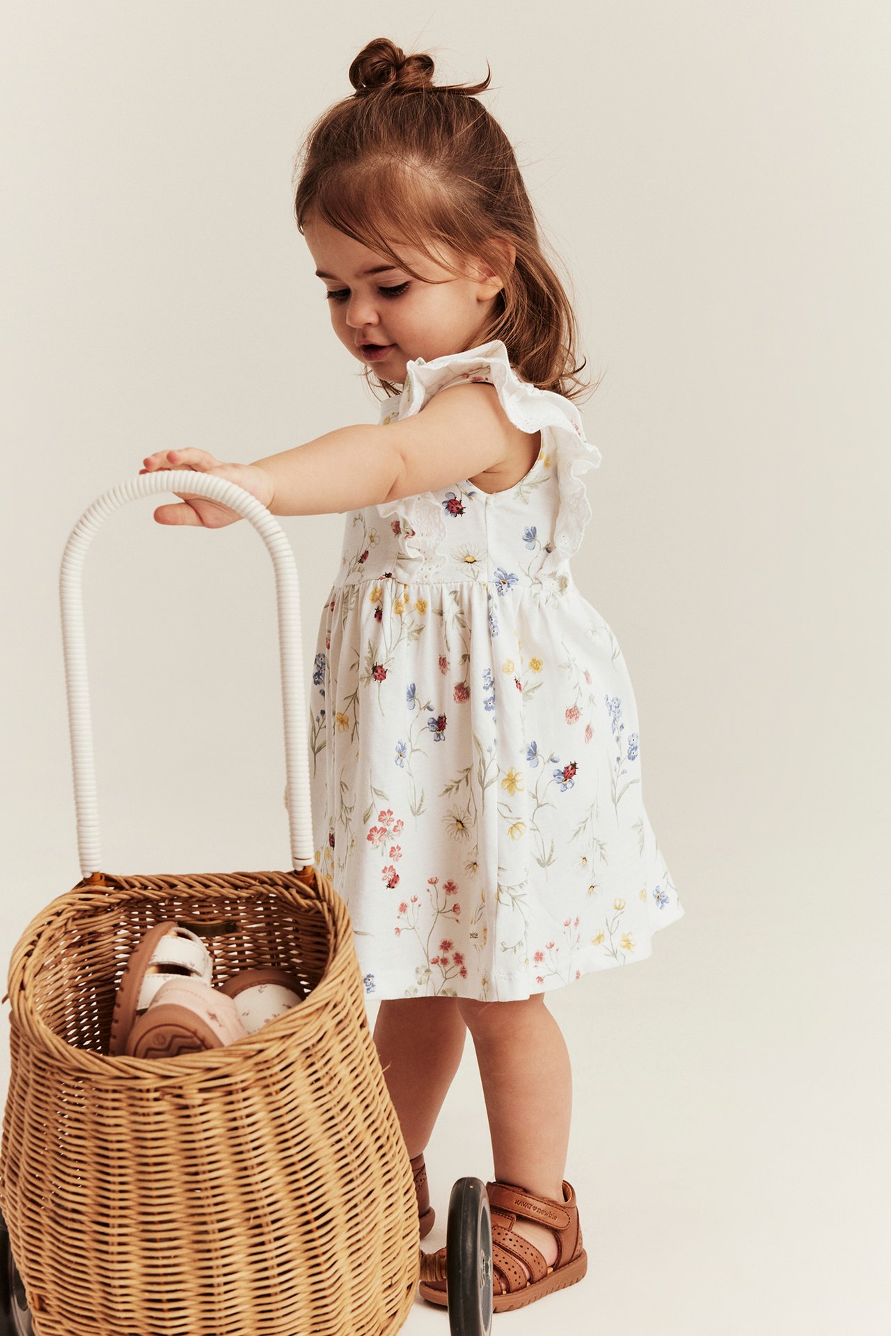 Sidebilde av baby i offwhite blomstret jerseykjole, volanger, brune sandaler, holder flettet vogn.