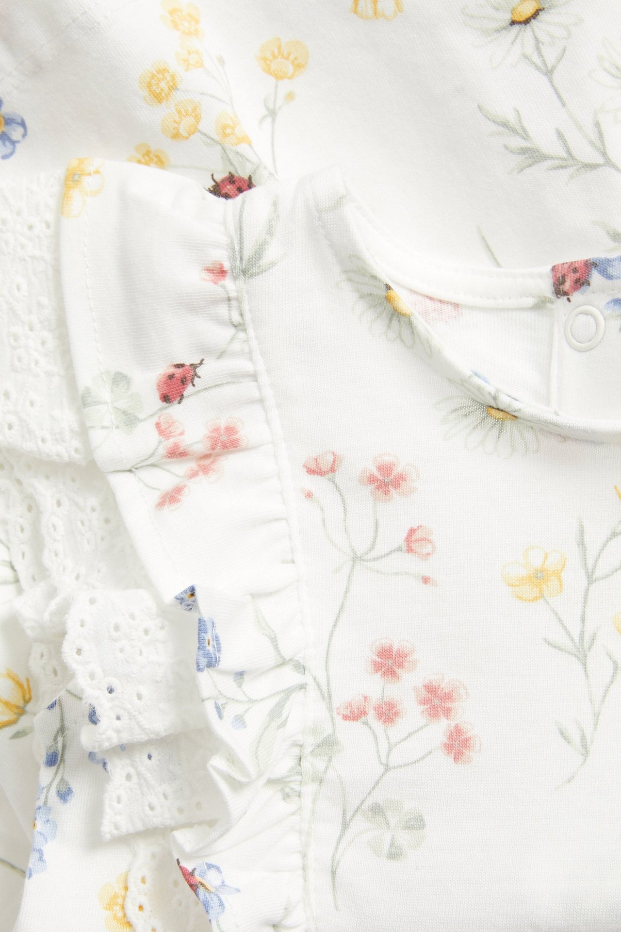 Närbild, framifrån, på en offwhite jerseyklänning i bomull med blommigt mönster, volanger och spetsdetaljer.