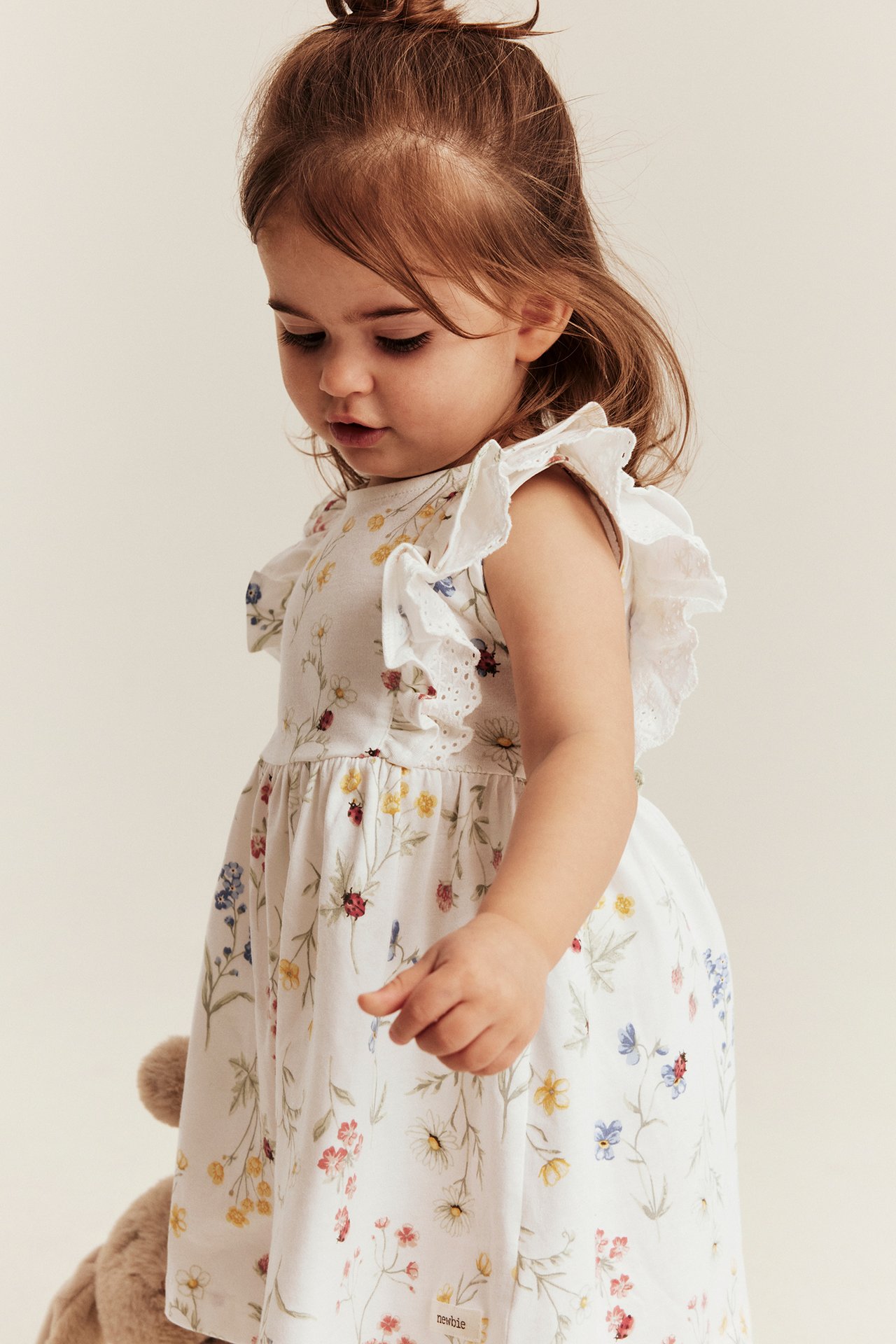 Widok z boku na niemowlę w ecru dżersejowej sukience w kwiaty, z falbankami i koronką.