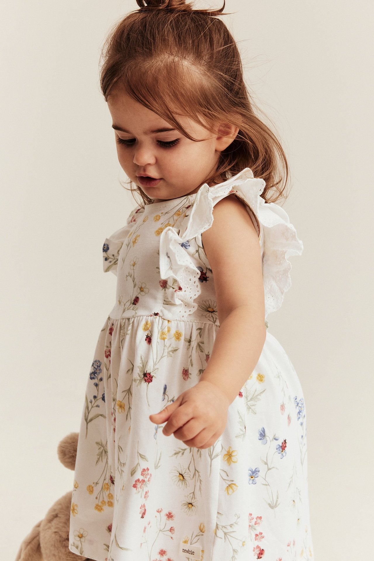 Widok z boku na niemowlę w ecru dżersejowej sukience w kwiaty, z falbankami i koronką.