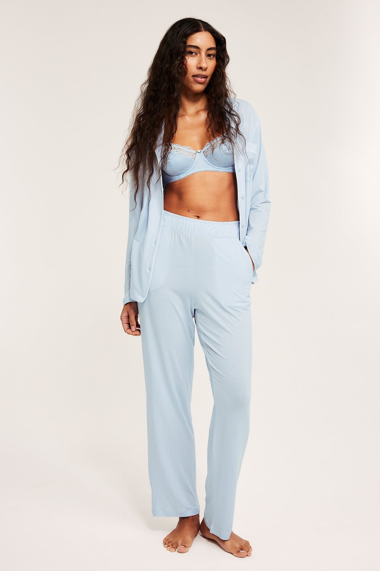 Widok z przodu: Kobieta w jasnoniebieskiej piżamie z lyocellu: rozpięta koszula, proste spodnie, koronkowy biustonosz.
