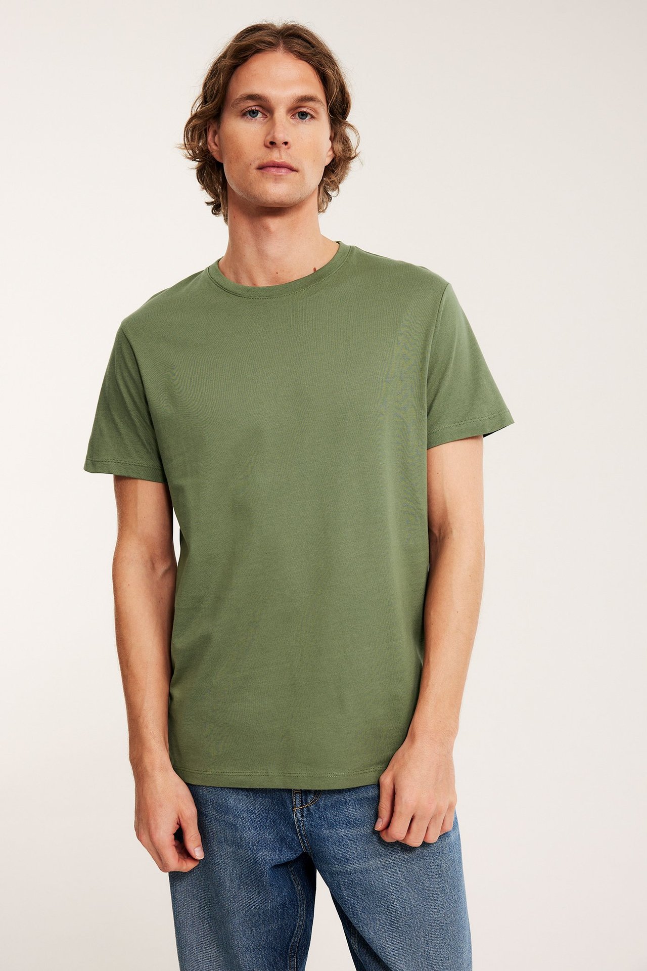 Edestä, mies yllään yksivärinen khakinvihreä pyöreäkauluksinen puuvilla-t-paita ja siniset farkut.