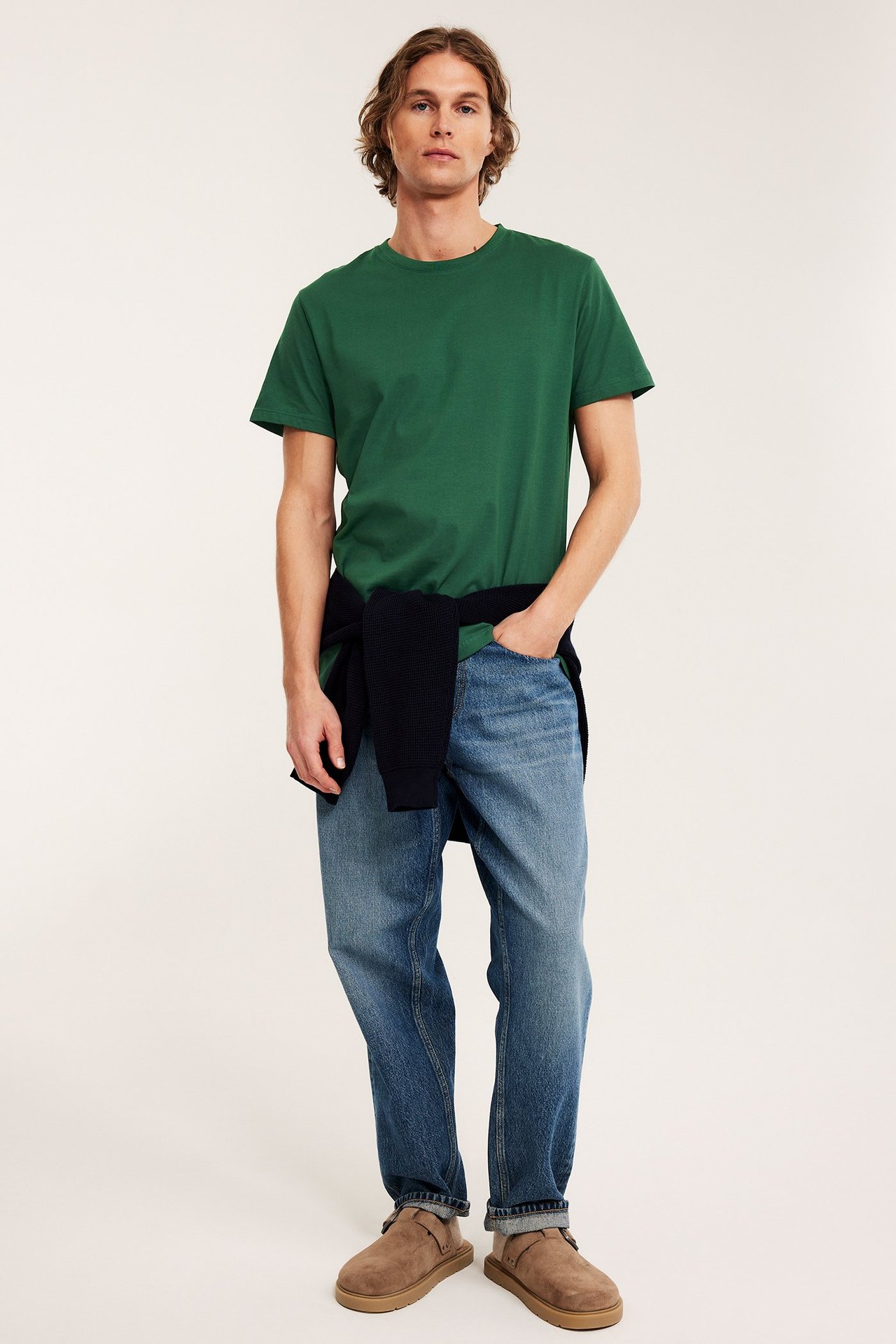 Mann i mørkegrønn t-skjorte, blå jeans, brune slip-on-sko og genser knyttet i midjen.