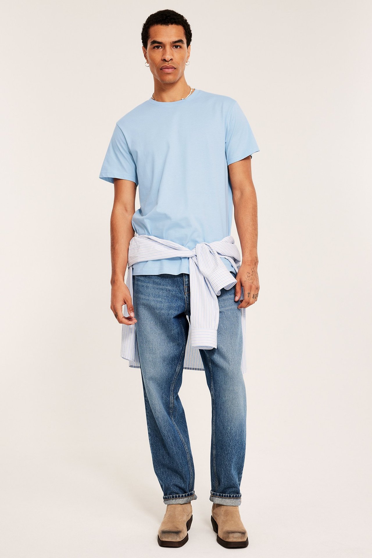 Mann i lyseblå t-skjorte, blå jeans, stripet skjorte i midjen, beige sko. Forfra.