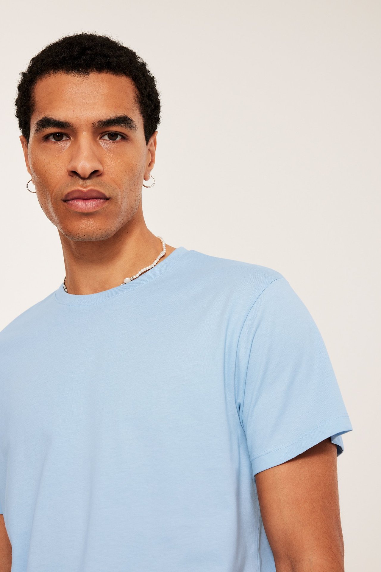 Zbliżenie mężczyzny w jasnoniebieskim, bawełnianym t-shircie z okrągłym dekoltem, naszyjniku z pereł i srebrnych kolczykach.