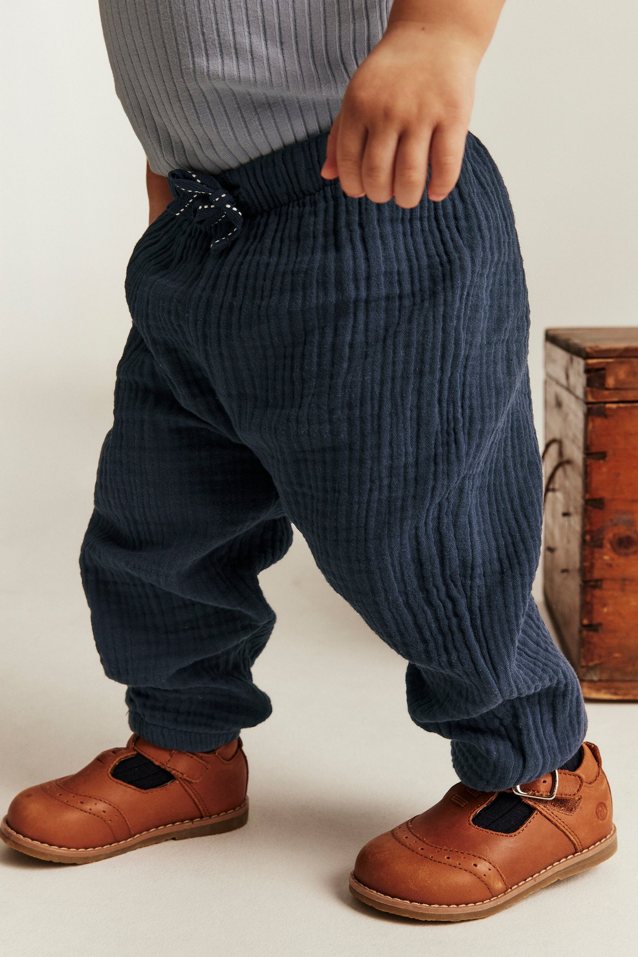 Etukuva: Vauva yllään tummansiniset kudotut musliinihousut joustovyötäröllä, harmaa ribattu toppi ja ruskeat nahkakengät.