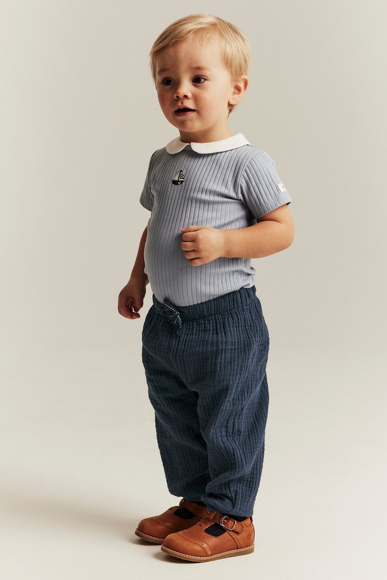 Chłopiec w niebieskich muślinowych spodniach ze sznurkiem, jasnoniebieskiej prążkowanej koszulce z kołnierzykiem i brązowych butach.
