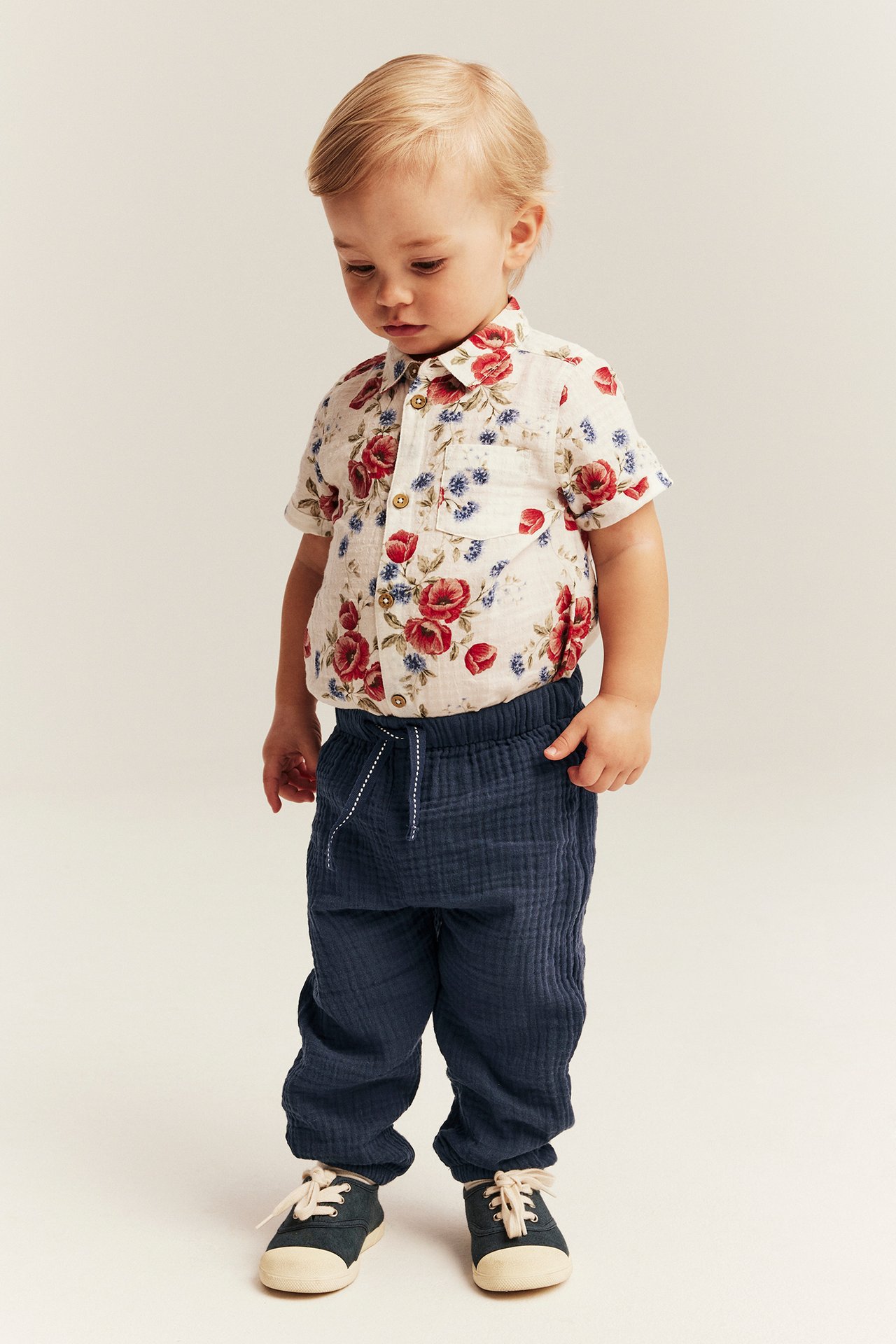 Frontvy: baby i mörkblå vävda muslinbyxor, en blommig skjorta och mörka sneakers.