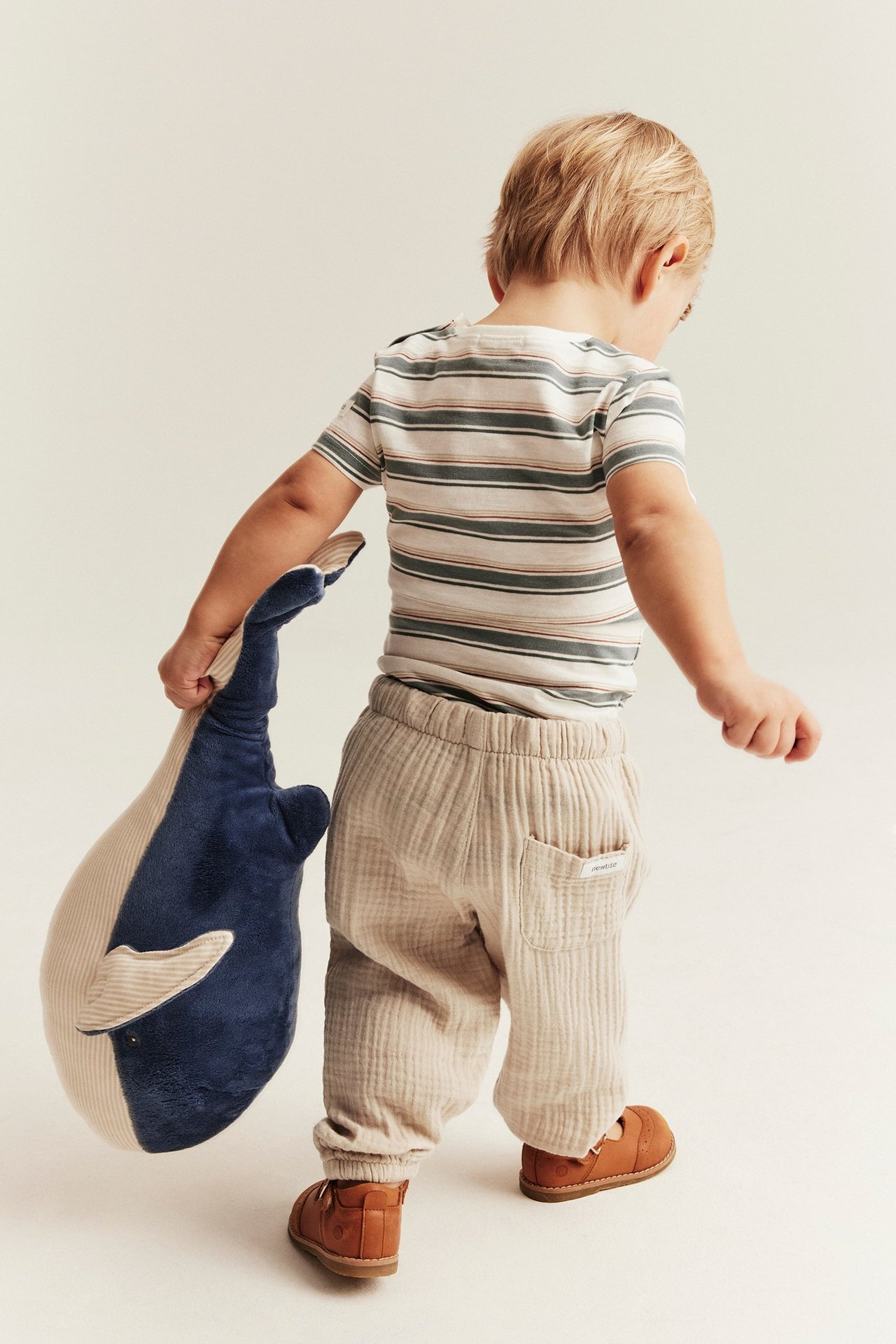 Ryggvy av ett barn i offwhite muslinbyxor, en randig t-shirt, bruna skor, håller en blå valmjukis.