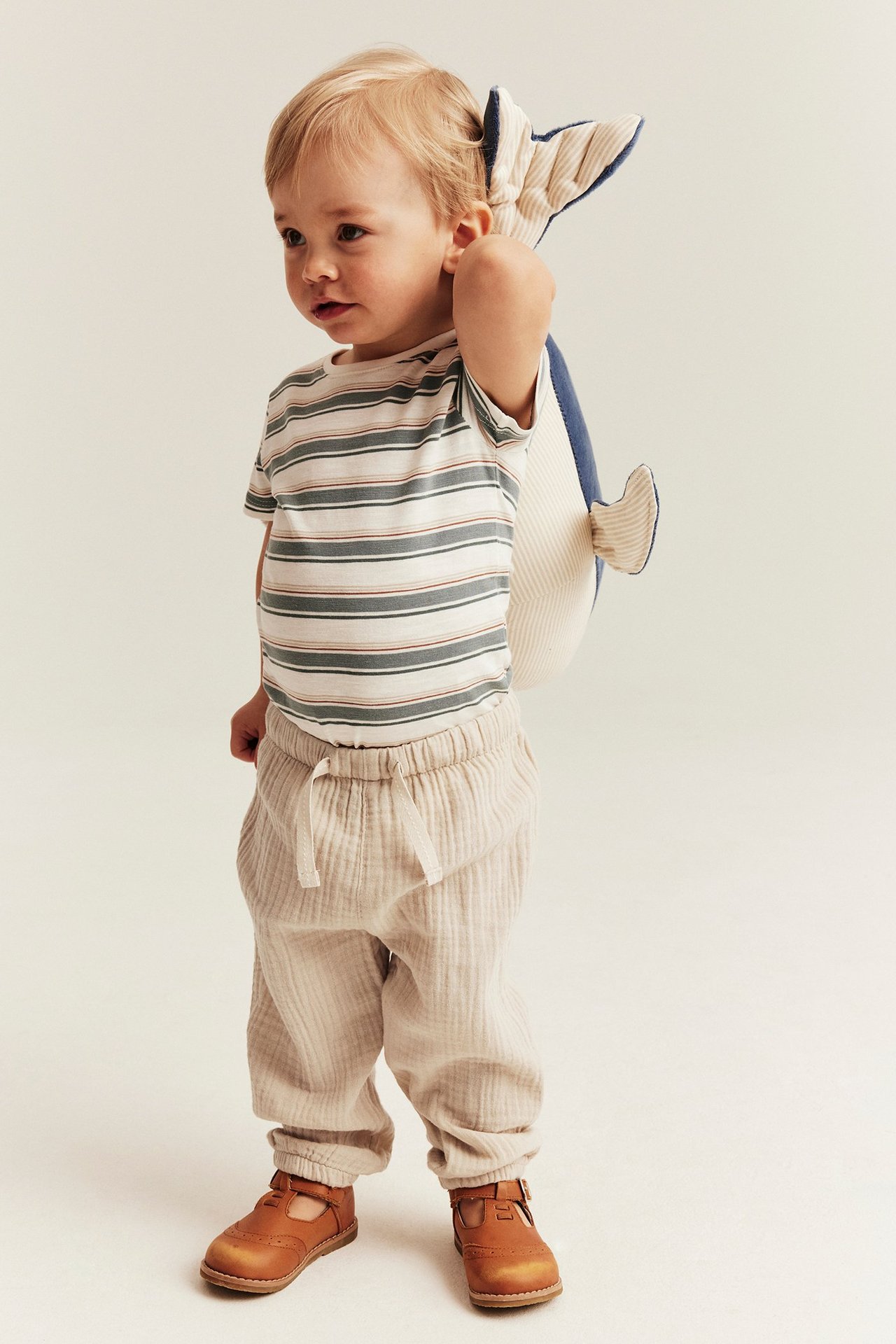 Forfra, baby i off-white vevde muslinbukser med strikk i midjen, stripet topp, brune sko, holder plysjhval.