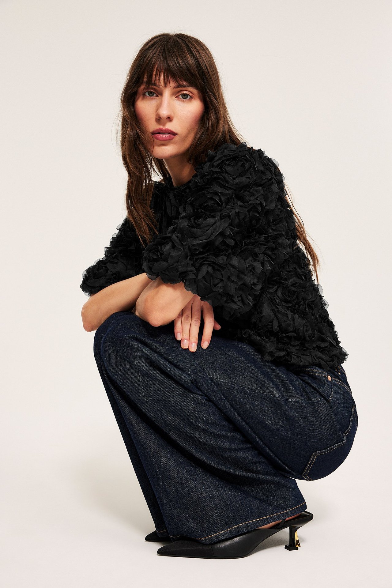 Kvinne i svart puffermbluse med knyting, mørke jeans, svarte hæler, front.