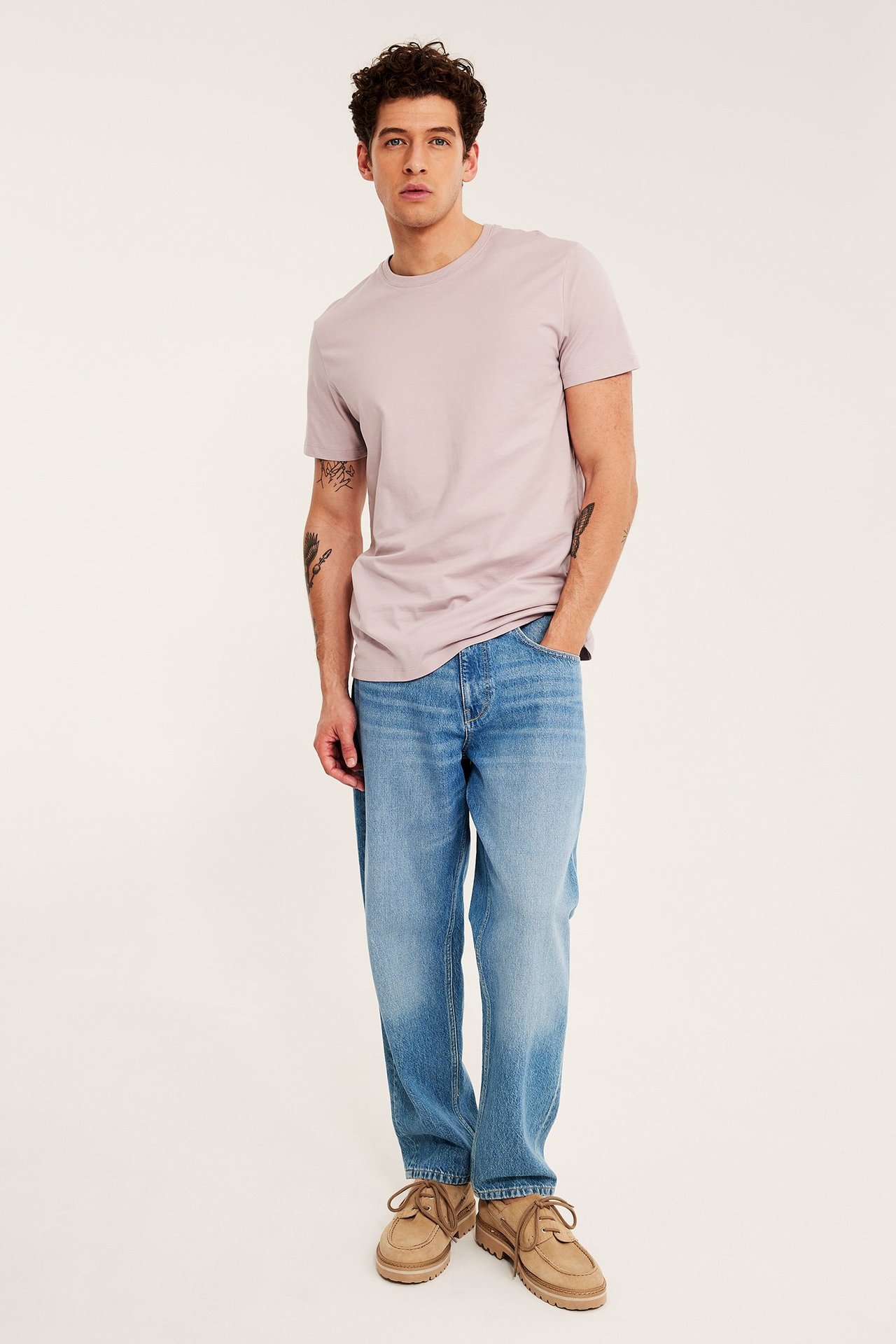 Framifrån en man i enfärgad rosa bomulls-t-shirt, blå jeans och ljusbruna snörskor.