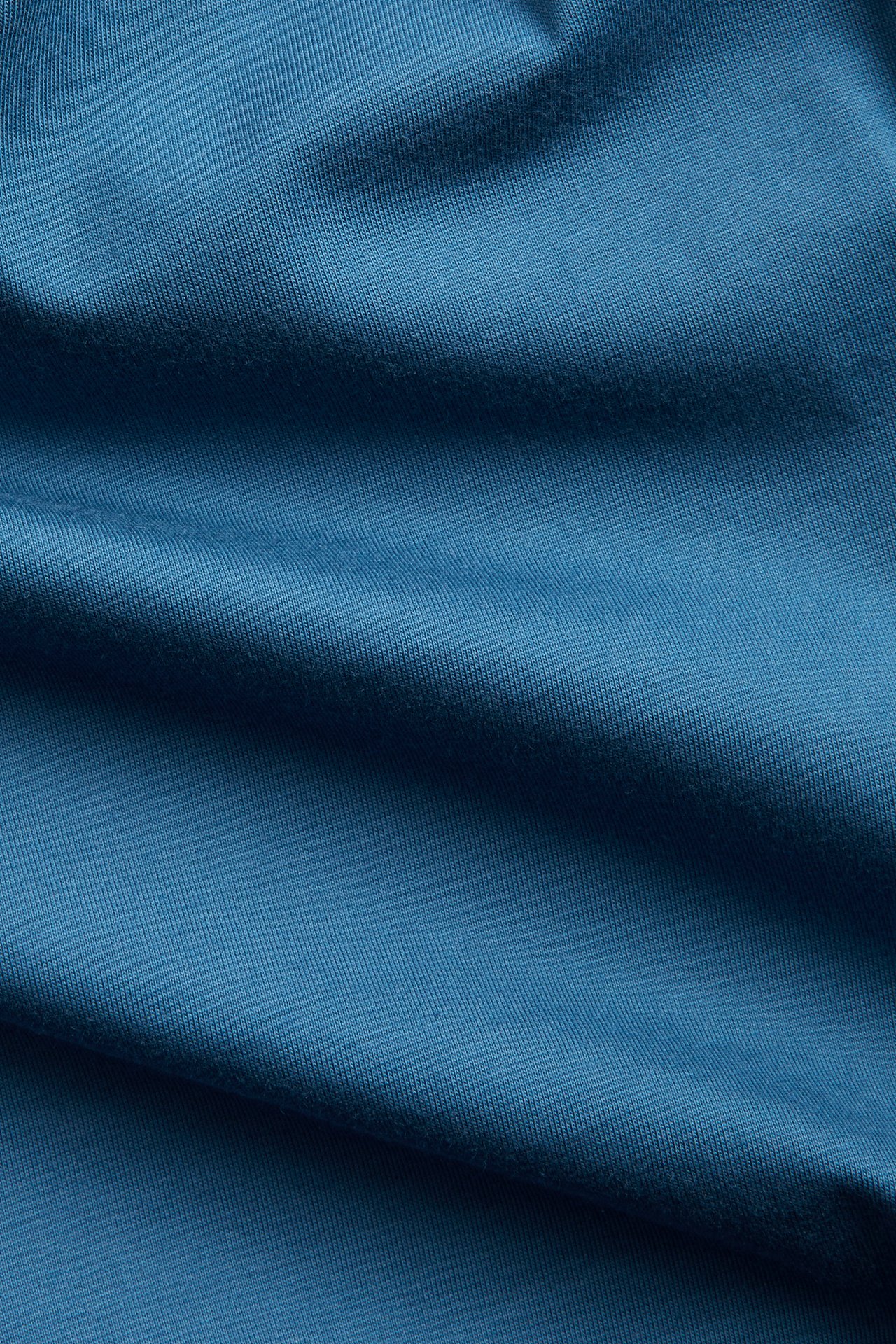Nærbilde av et blått, ensfarget bomullsstoff, typisk for en rundhalset herre-T-skjorte.
