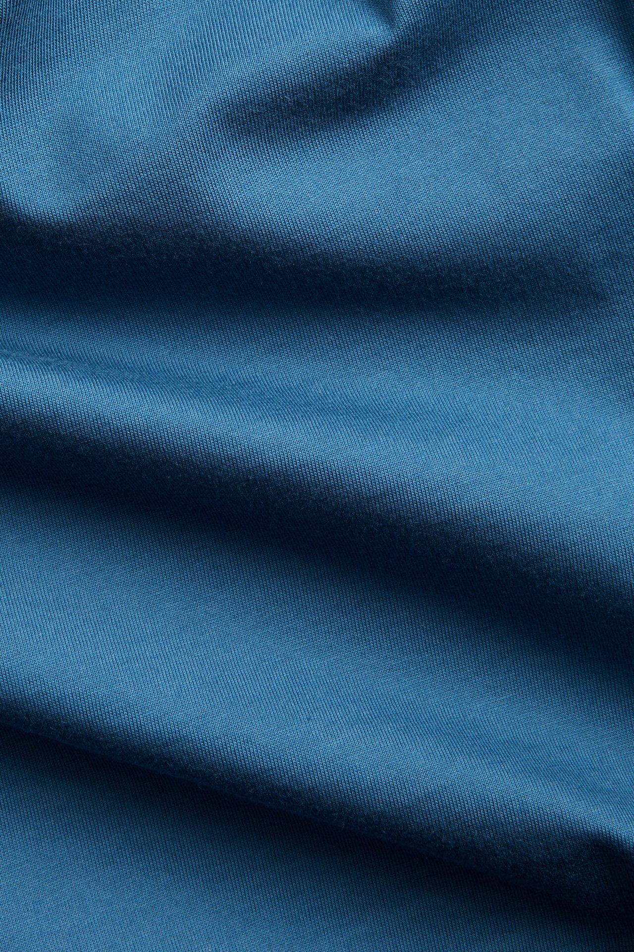 Nærbilde av et blått, ensfarget bomullsstoff, typisk for en rundhalset herre-T-skjorte.
