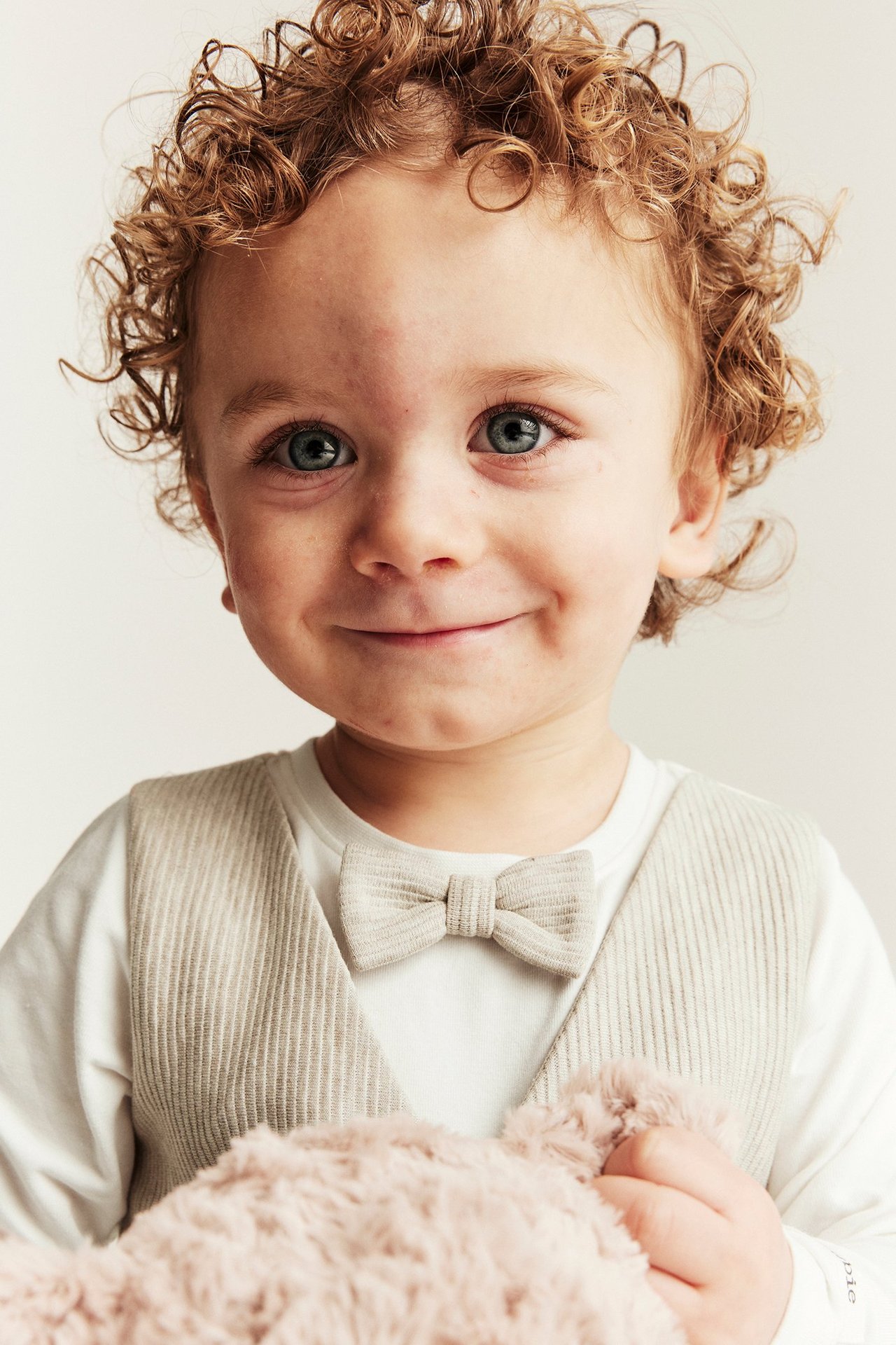 Frontvisning: Smilende baby i beige melert body med påsydd vest og sløyfe, holder et rosa plysjdyr.