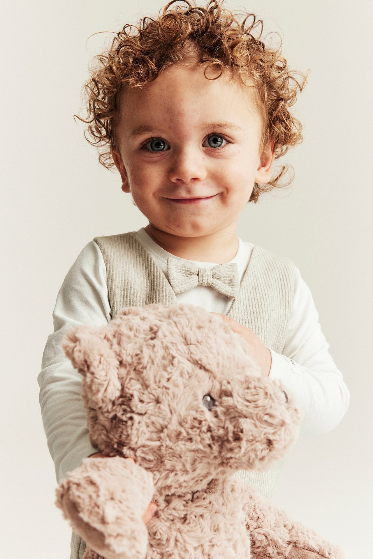Forfra: Smilende baby har på seg en beige vestbody med sløyfe, holder en myk brun bamse.