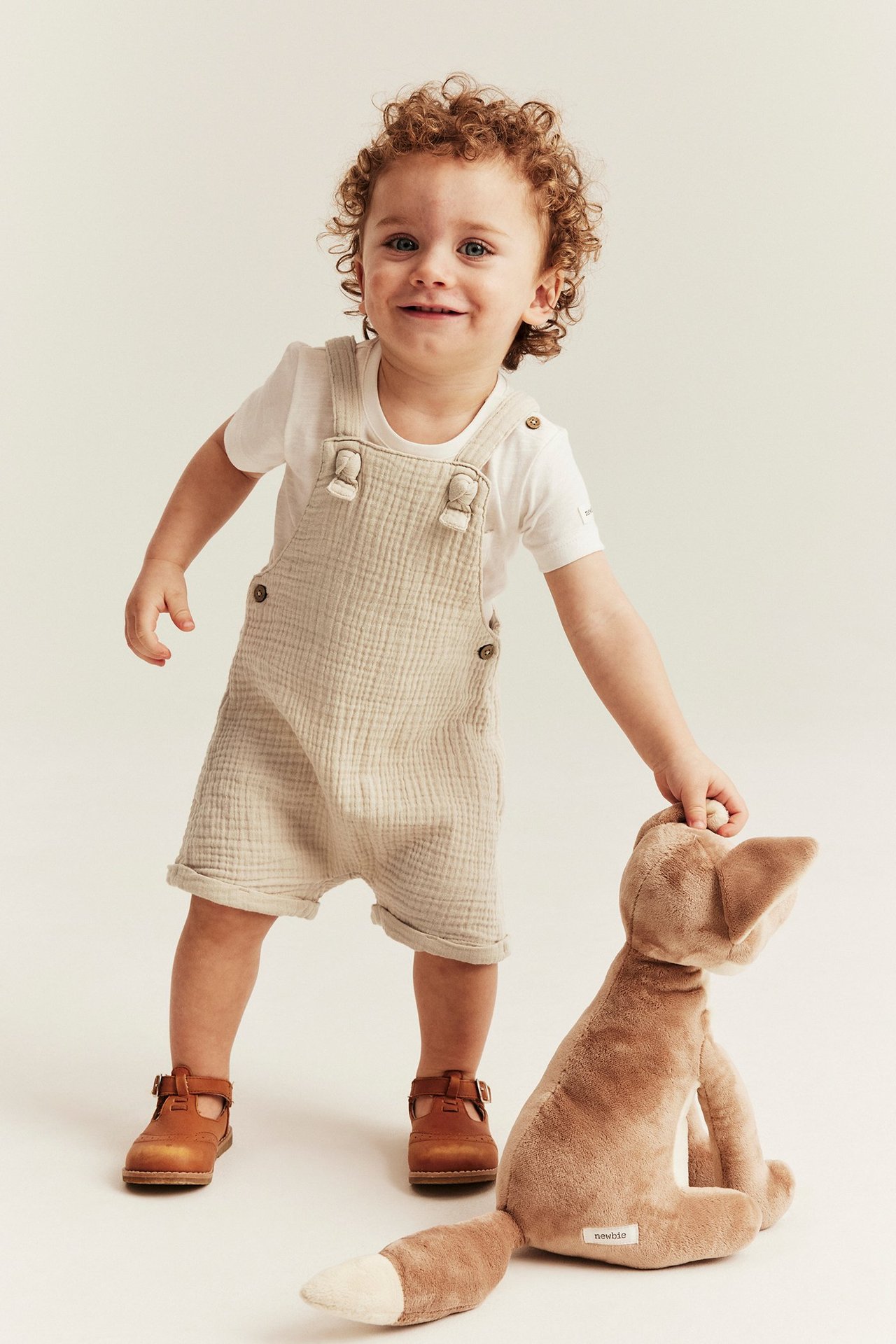 Edestä: Vauva yllään luonnonvalkoiset musliinihaalarihousut, valkoinen t-paita ja ruskeat kengät, pitäen pehmokettua.