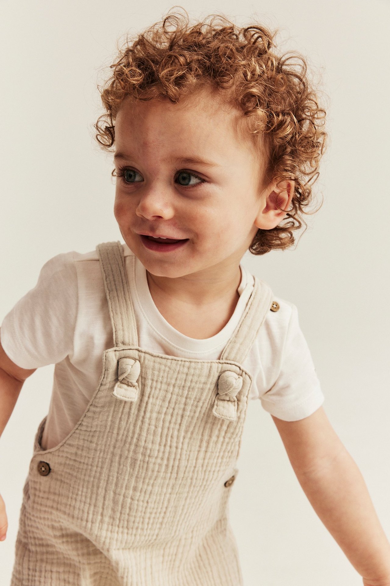 Närbild, framifrån: baby i offwhite vävda hängselshorts i muslin med knutna hängslen över en vit t-shirt.