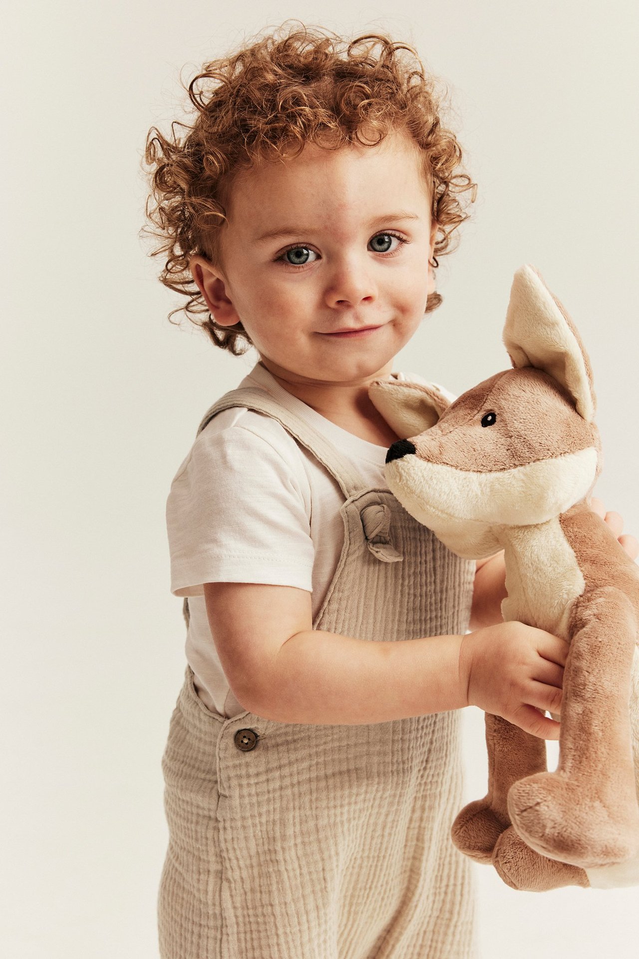 Edestäpäin vauva, yllään luonnonvalkoiset kudotut musliinihaalarishortsit, alla luonnonvalkoinen t-paita, pitäen ruskeaa pehmolelua.