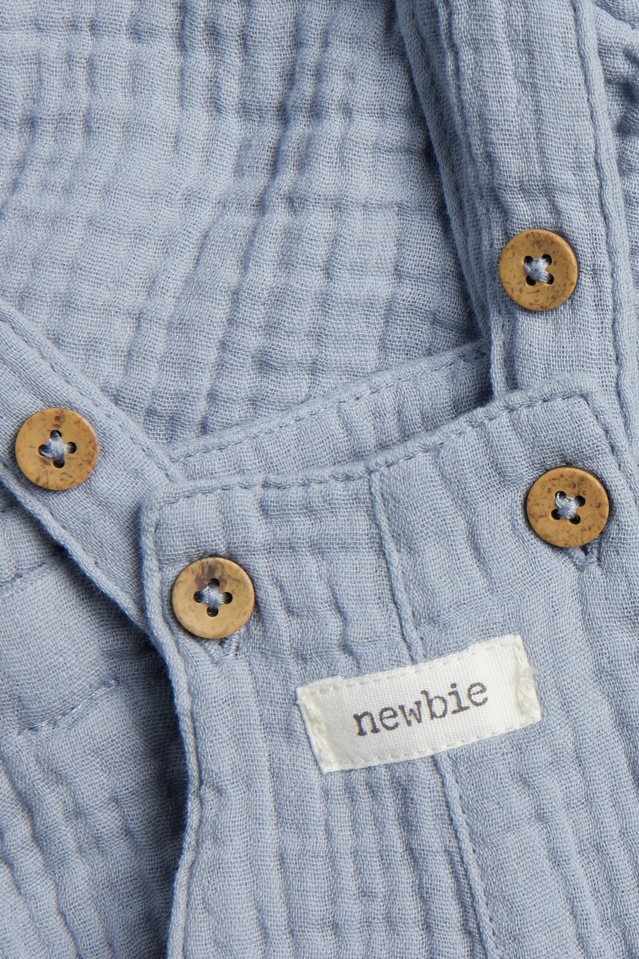 Zbliżenie: jasnoniebieskie muślinowe szorty ogrodniczki z drewnianymi guzikami i metką Newbie.