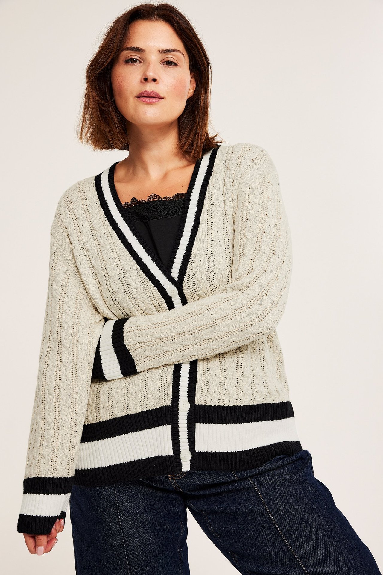 Kvinne i offwhite flettet V-hals cardigan med svart/hvite striper, over svart blonde-topp og mørke jeans.