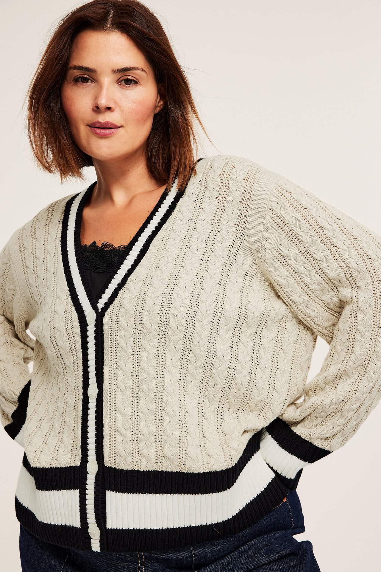 Forfra, kvinne i offwhite flettestrikket bomullscardigan med svart/hvit stripet kant over svart topp.