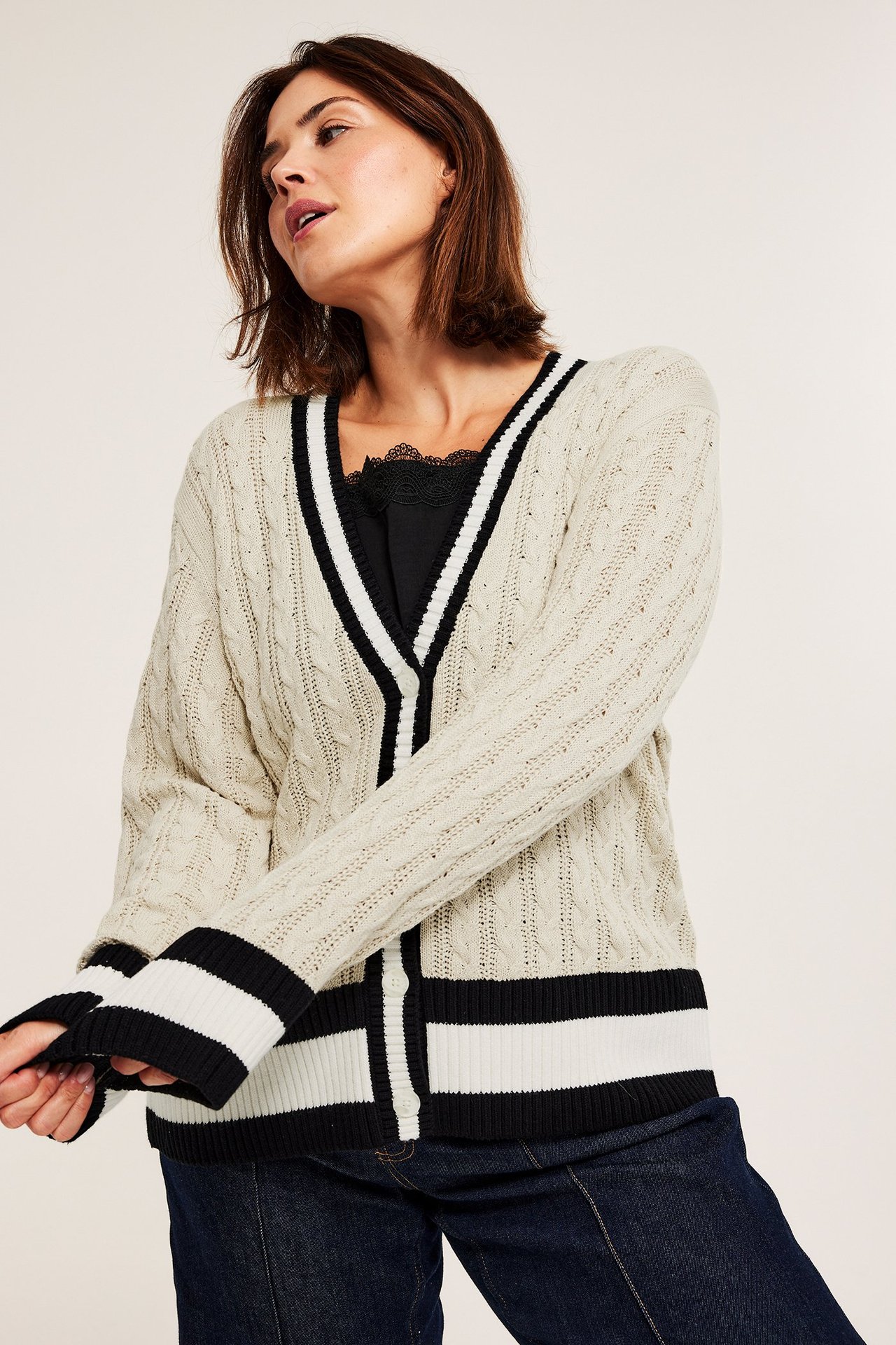 Kvinne: Kremfarget kabelstrikket cardigan med stripedetaljer, svart blonde topp, mørkeblå jeans.