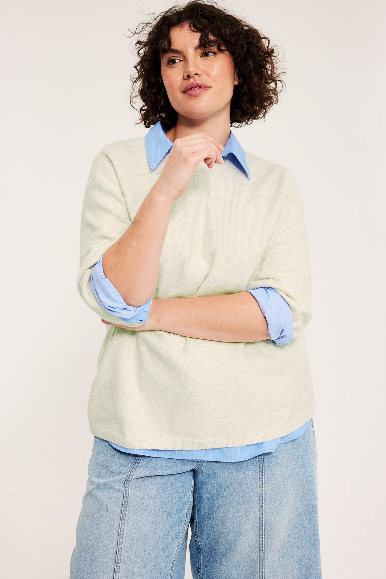 Widok z przodu: Kobieta w kremowym melanżowym swetrze 3/4 rękawa na niebieskiej koszuli w paski i jasnych jeansach.