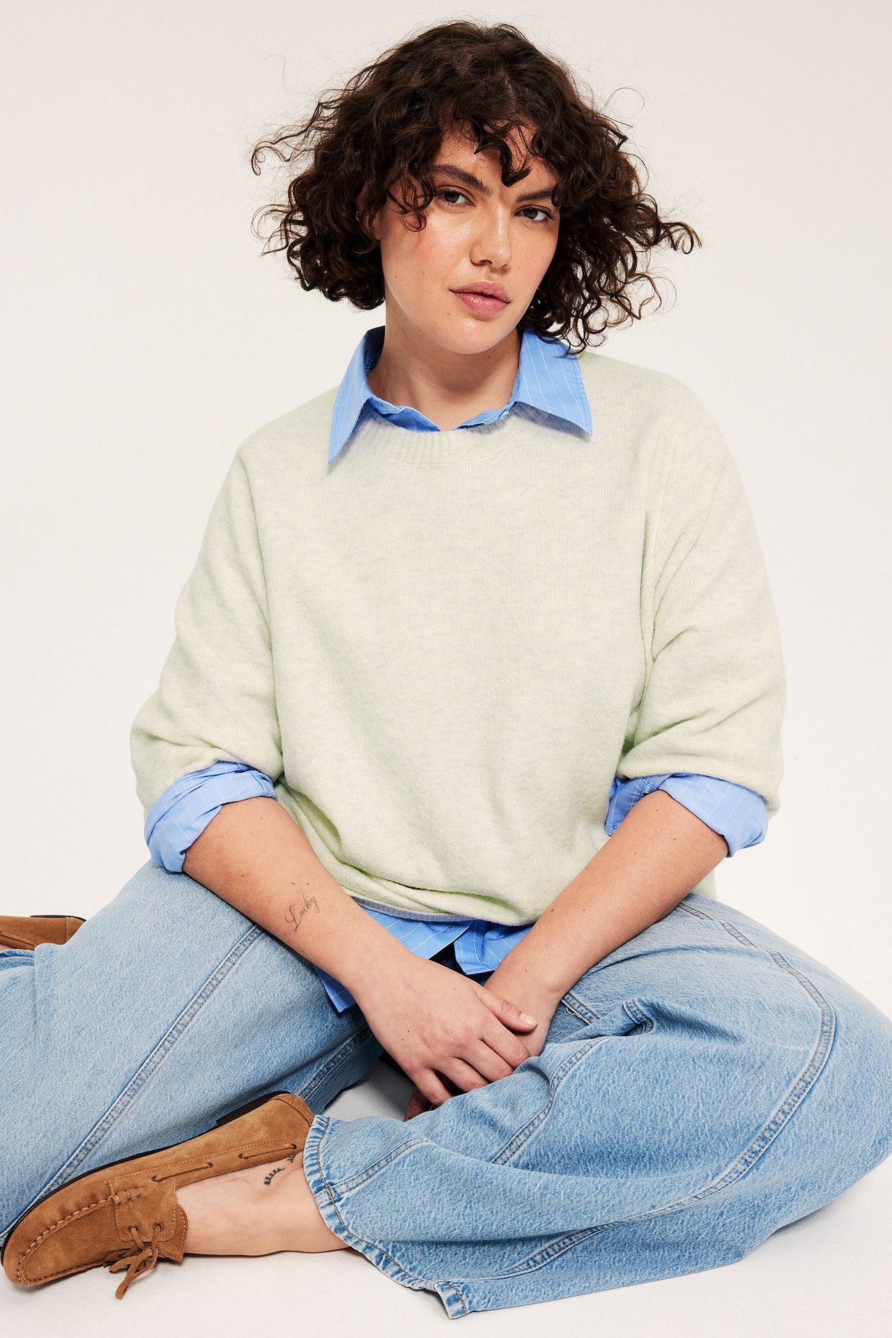 Widok z przodu: Kobieta w ecru melanżowej dzianinowej koszulce 3/4, niebieskiej koszuli, jasnych jeansach, brązowych mokasynach.