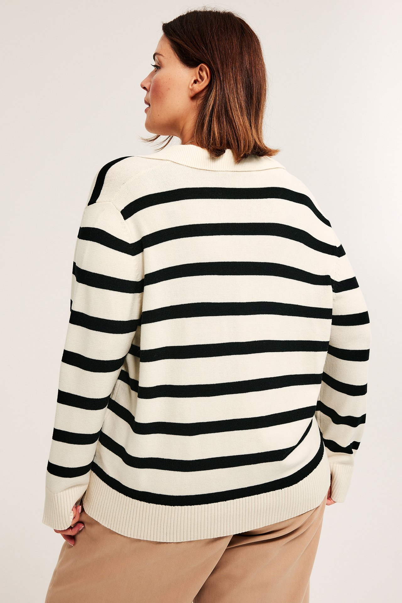 Høyre baksidevisning av en kvinne i en offwhite- og svartstripet strikket genser med krage og brune bukser.