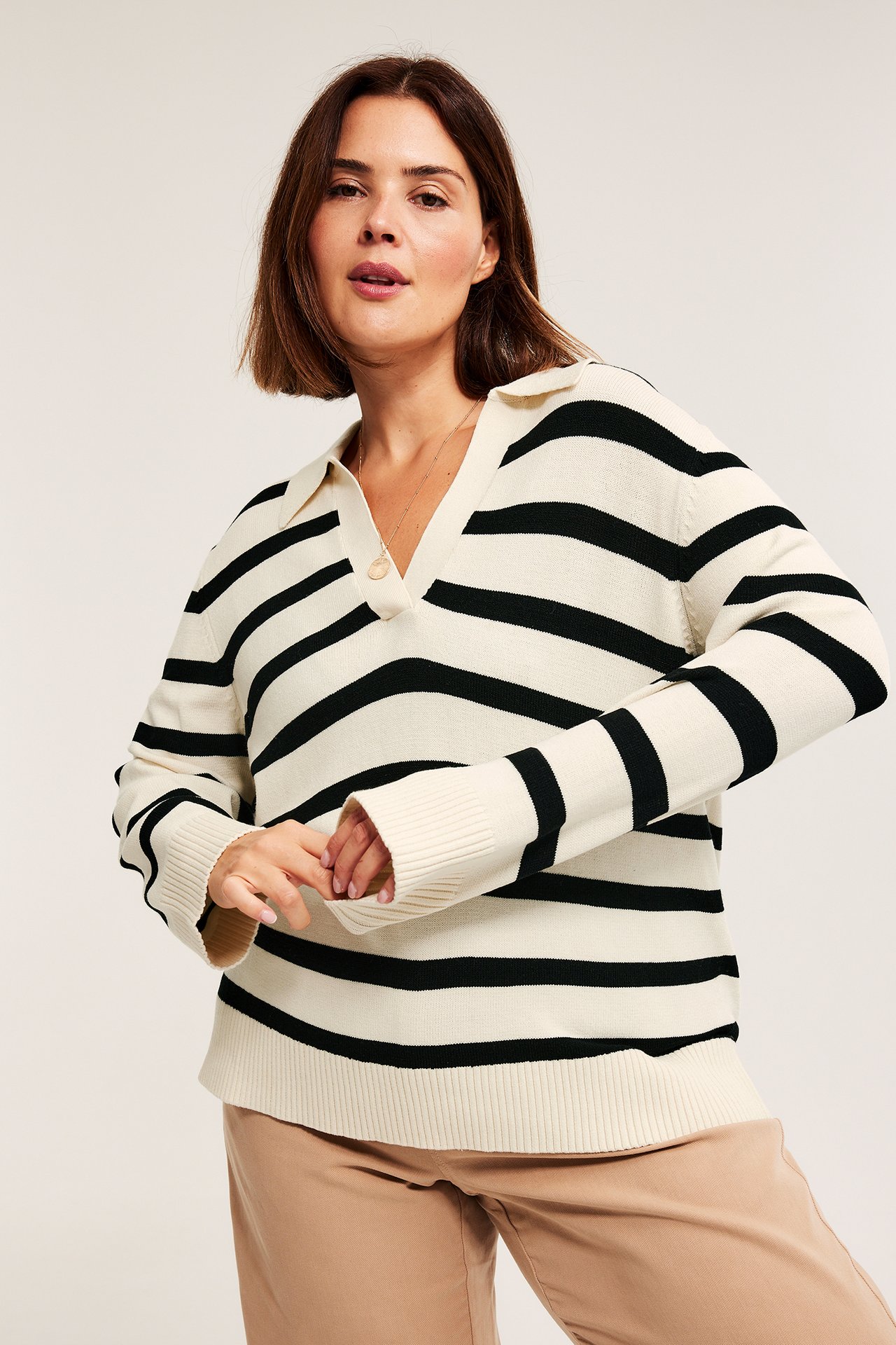Frontvisning av kvinne i løs offwhite, svartstripet strikkegenser med krage, v-hals, og beige bukse.
