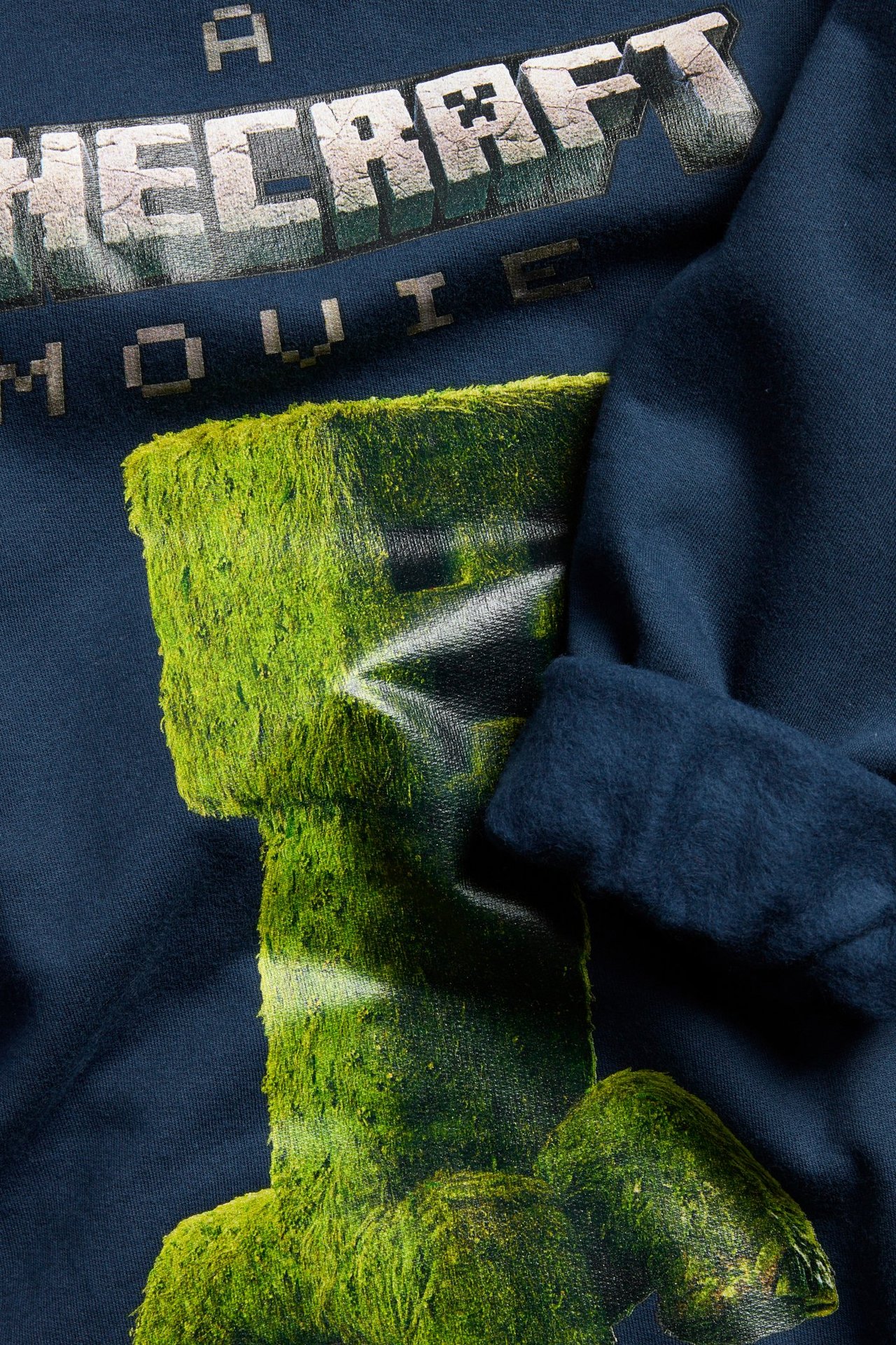 Nærbilde av en mørkeblå bomullssweatshirt med børstet innside og grønt Minecraft Movie-trykk.