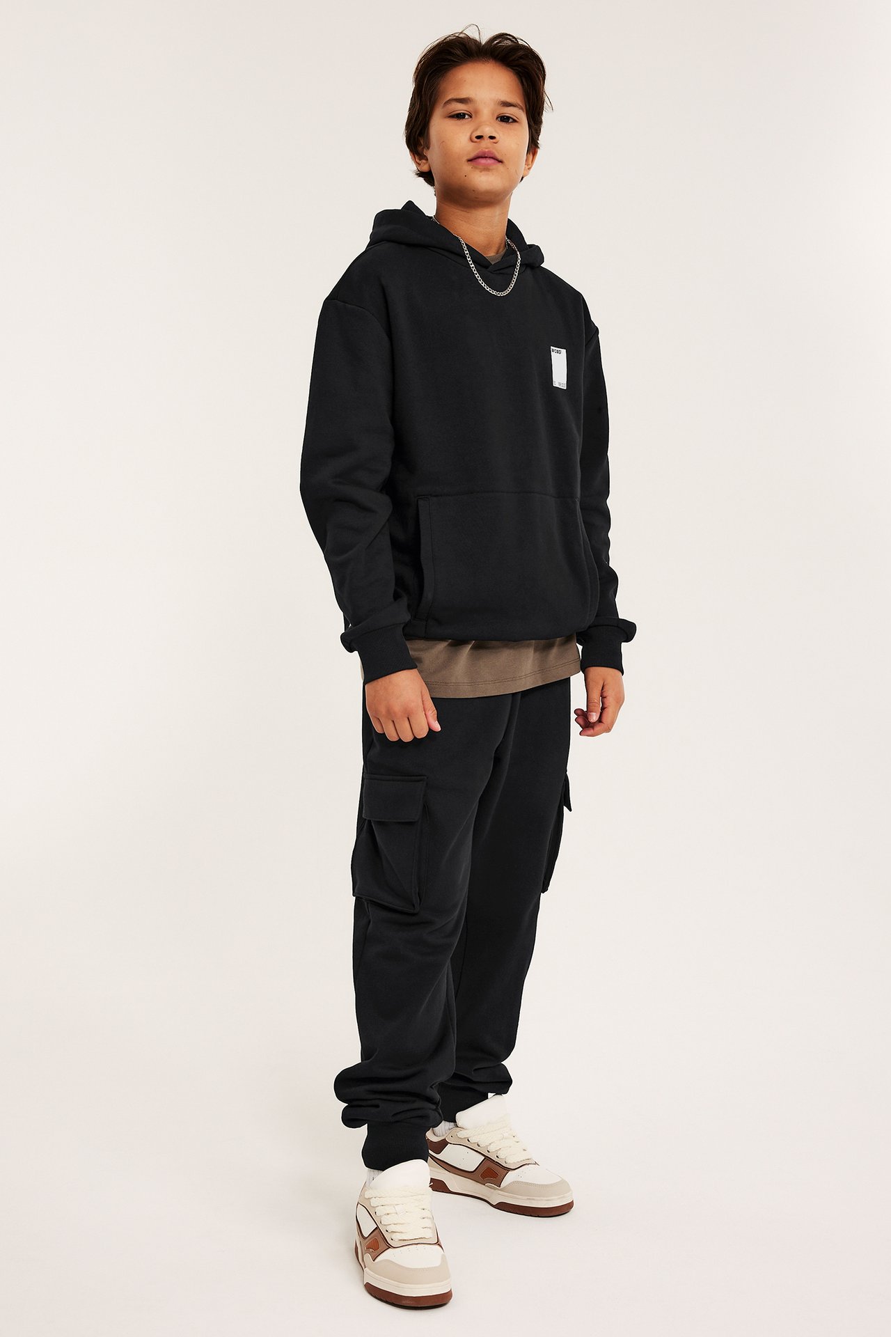 Widok z przodu i z boku: dziecko w czarnej bluzie z nadrukiem, czarnych bojówkach, brązowym t-shircie i sneakersach.