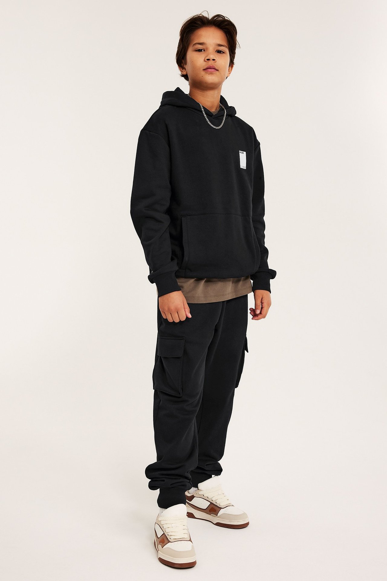 Edestä ja sivulta kuvattu lapsi mustassa painetussa hupparissa, mustissa cargohousuissa, ruskeassa t-paidassa ja lenkkareissa.