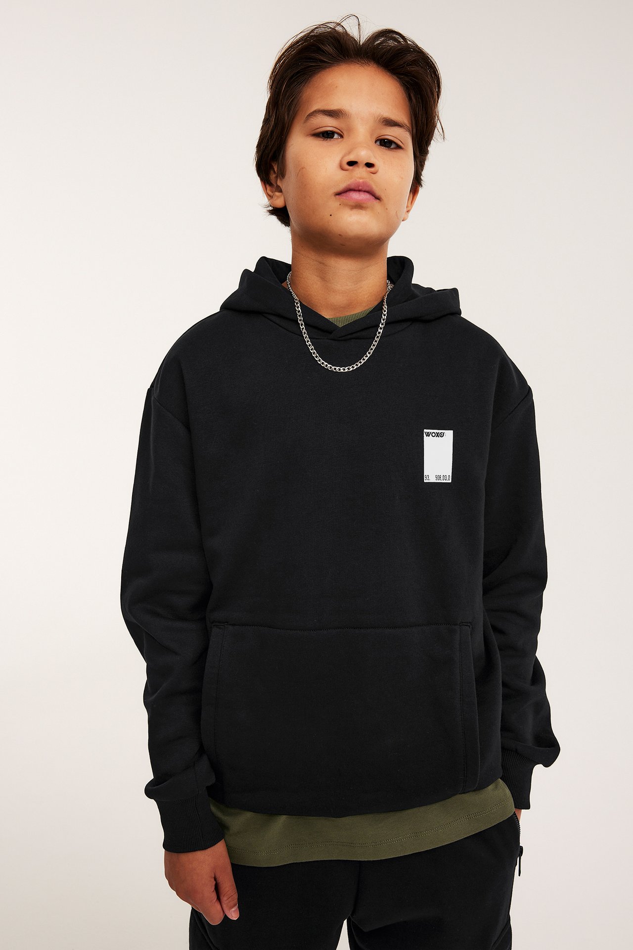 Framifrån, en kille bär en svart huvtröja med framfickor, vitt brösttryck, grön t-shirt och silverkedja.