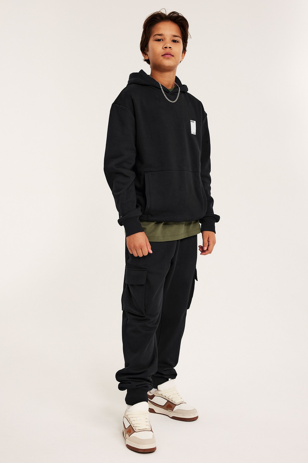 Widok z przodu, dziecko w czarnej bluzie z kapturem z nadrukiem, oliwkowym t-shircie, czarnych bojówkach i beżowych sneakersach.