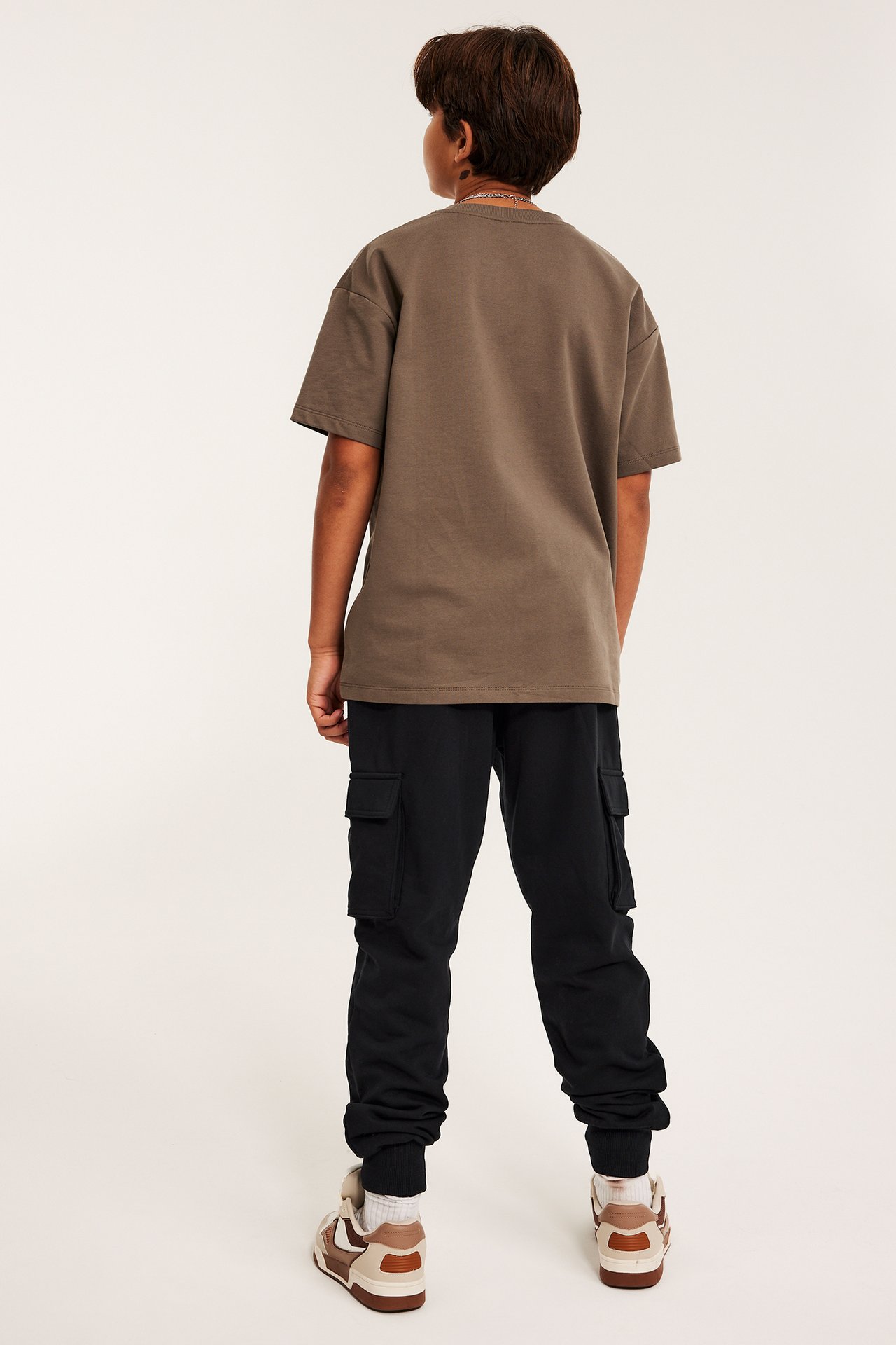 Widok z tyłu dziecka w czarnych spodniach dresowych z kieszeniami cargo, brązowej koszulce i brązowo-beżowych sneakersach.