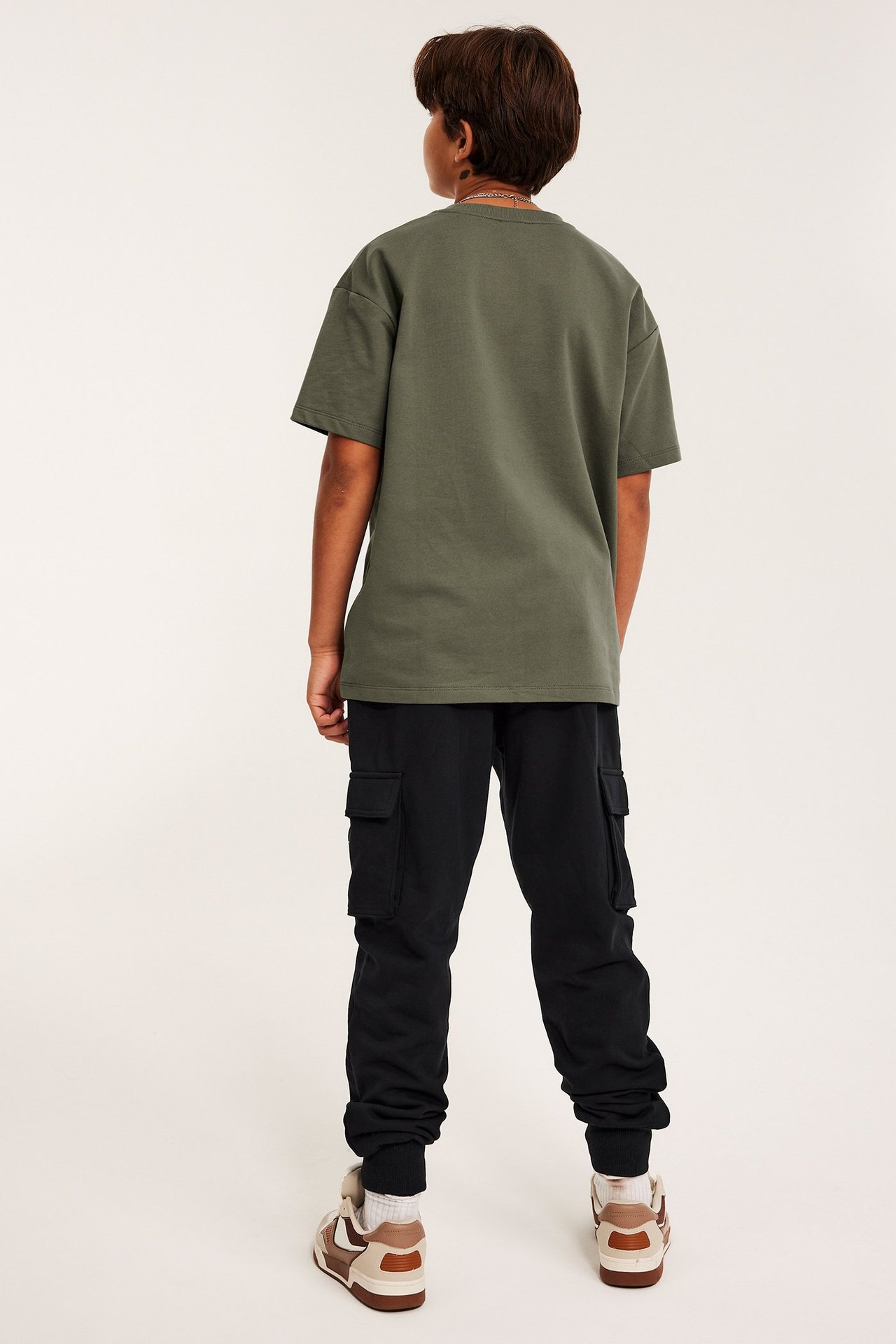 Bakifrån: ett barn i svarta mjukisbyxor med cargofickor, en olivgrön t-shirt och bruna sneakers.