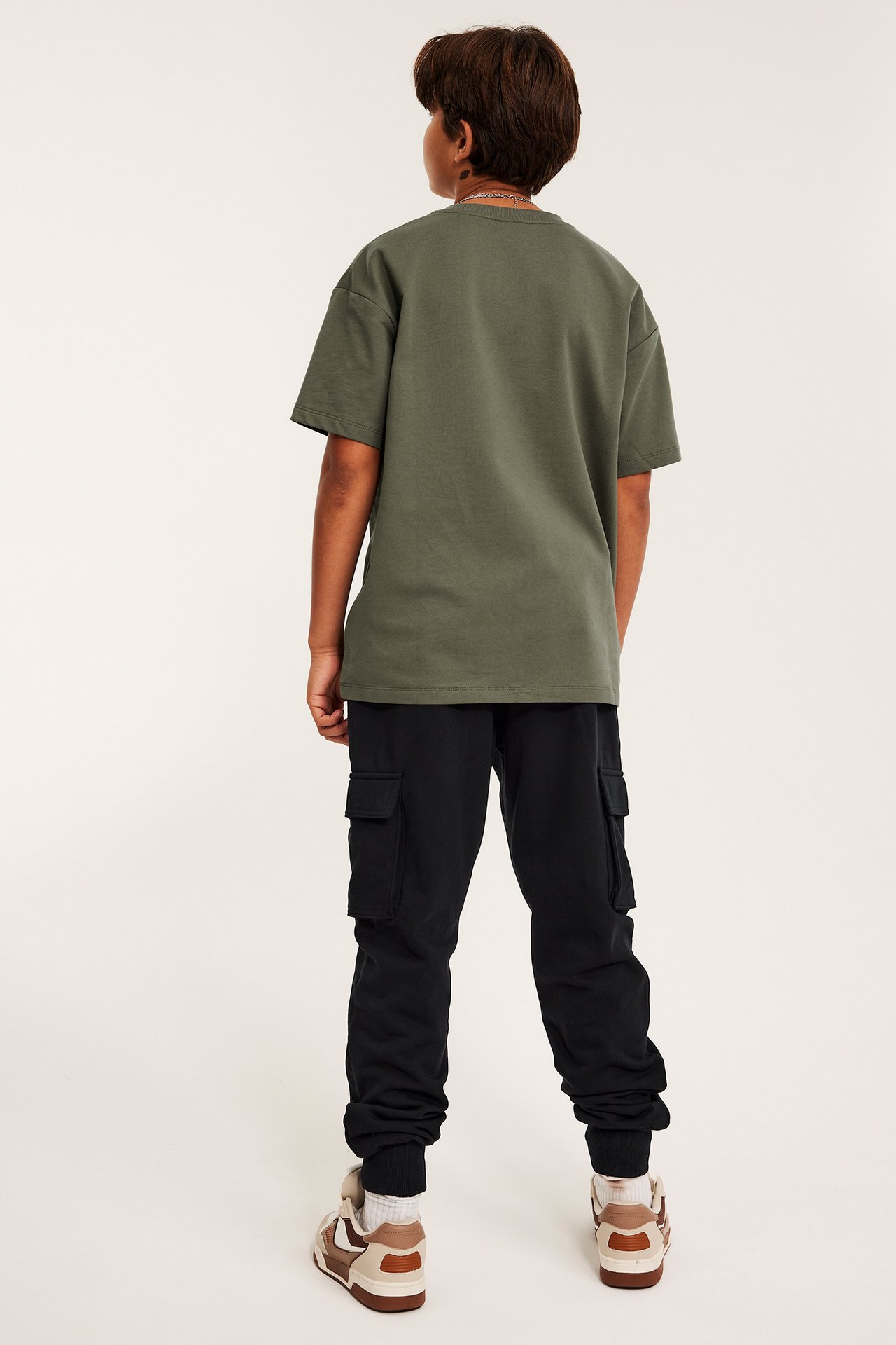 Bakifrån: ett barn i svarta mjukisbyxor med cargofickor, en olivgrön t-shirt och bruna sneakers.
