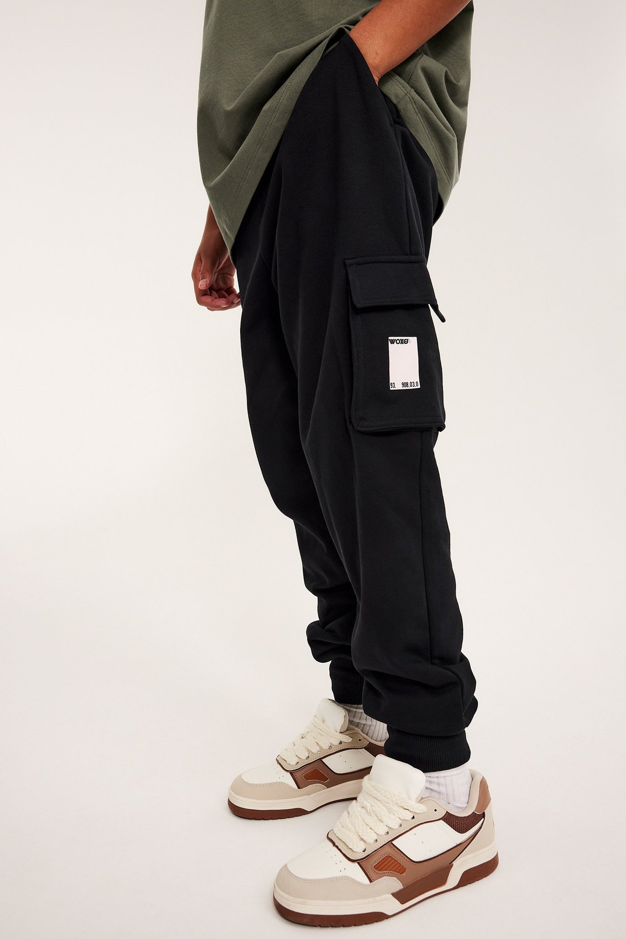 Lapsi yllään mustat collegehousut rahtitaskuilla ja painatuksella, oliivinvihreä t-paita ja valkoinen-ruskeat lenkkarit.